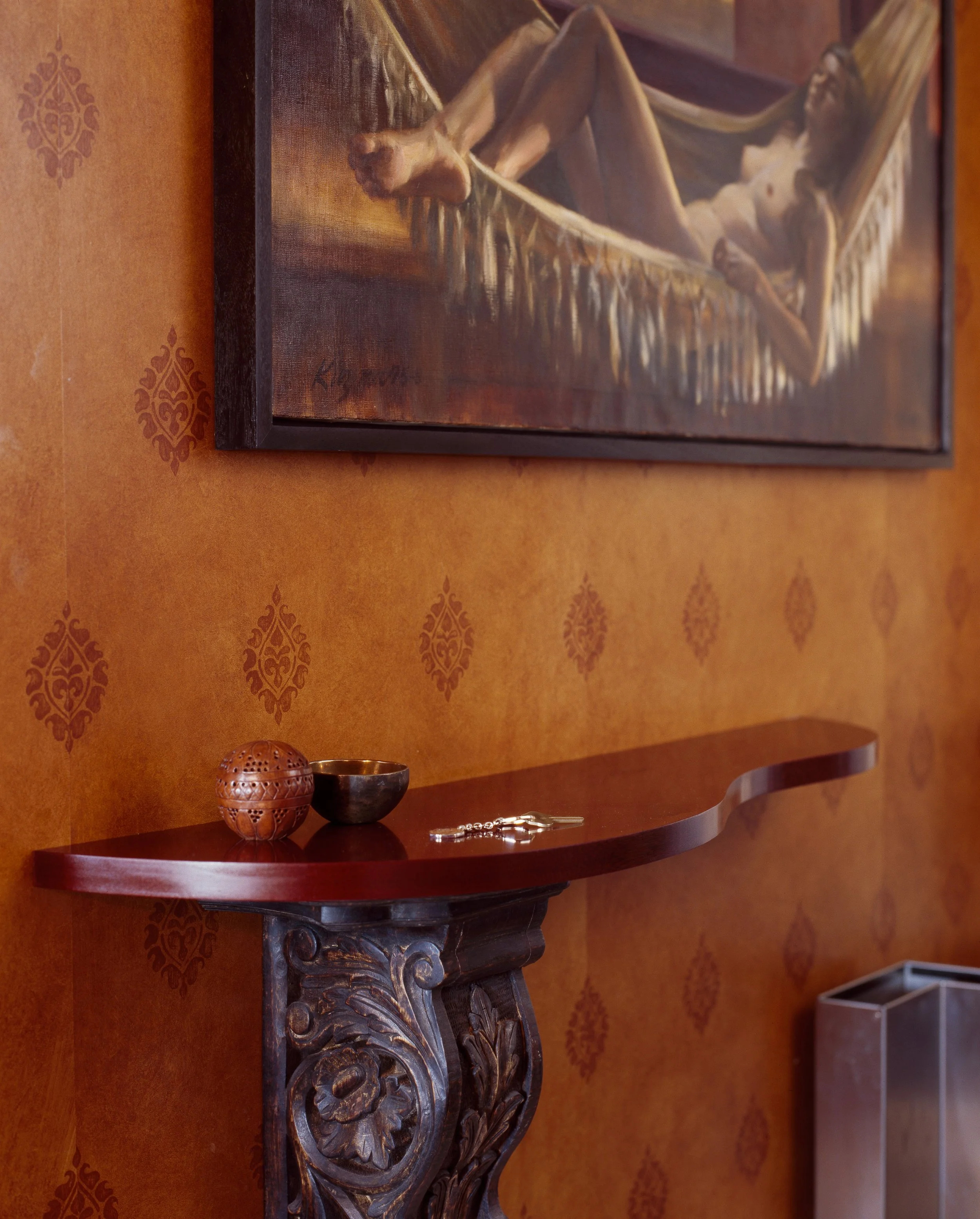 Close-up of a corner of a wall with a painting of a reclining nude woman on the wall. The painting is signed in the bottom left corner. Below the painting is a wooden console table with a carved ornamental support, on which there are two decorative objects: a carved round box and a small bowl. The background wall has orange textured wallpaper with a subtle pattern.