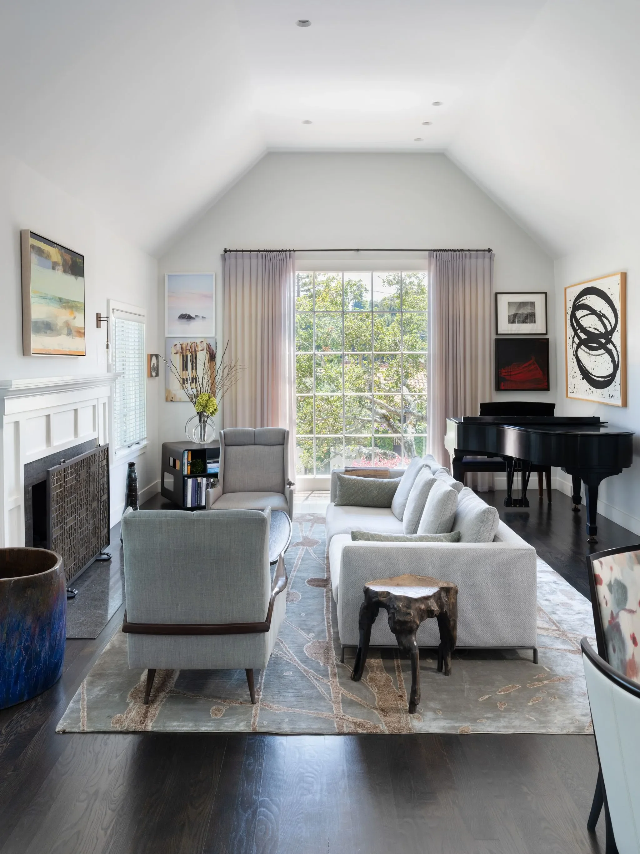Living room with large window, white walls, framed artwork, a black grand piano, and beige seating. There is a floral arrangement, a colorful ceramic vase, and a rustic wooden side table.