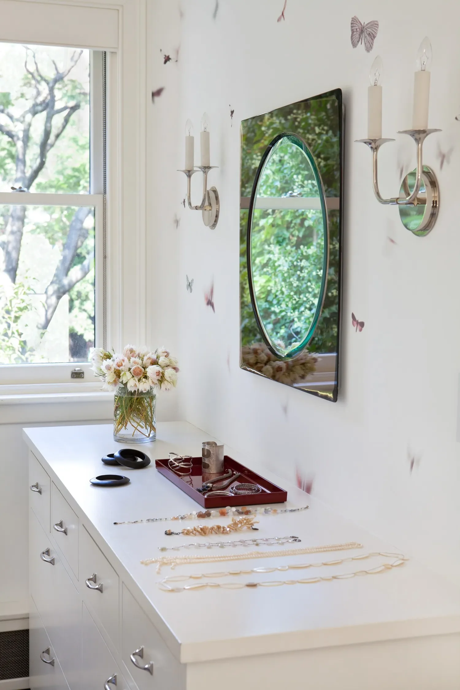 A white dresser with jewelry and accessories, a glass vase with pink and white flowers, and a mirror with a black oval frame on a white wall decorated with butterfly stickers.