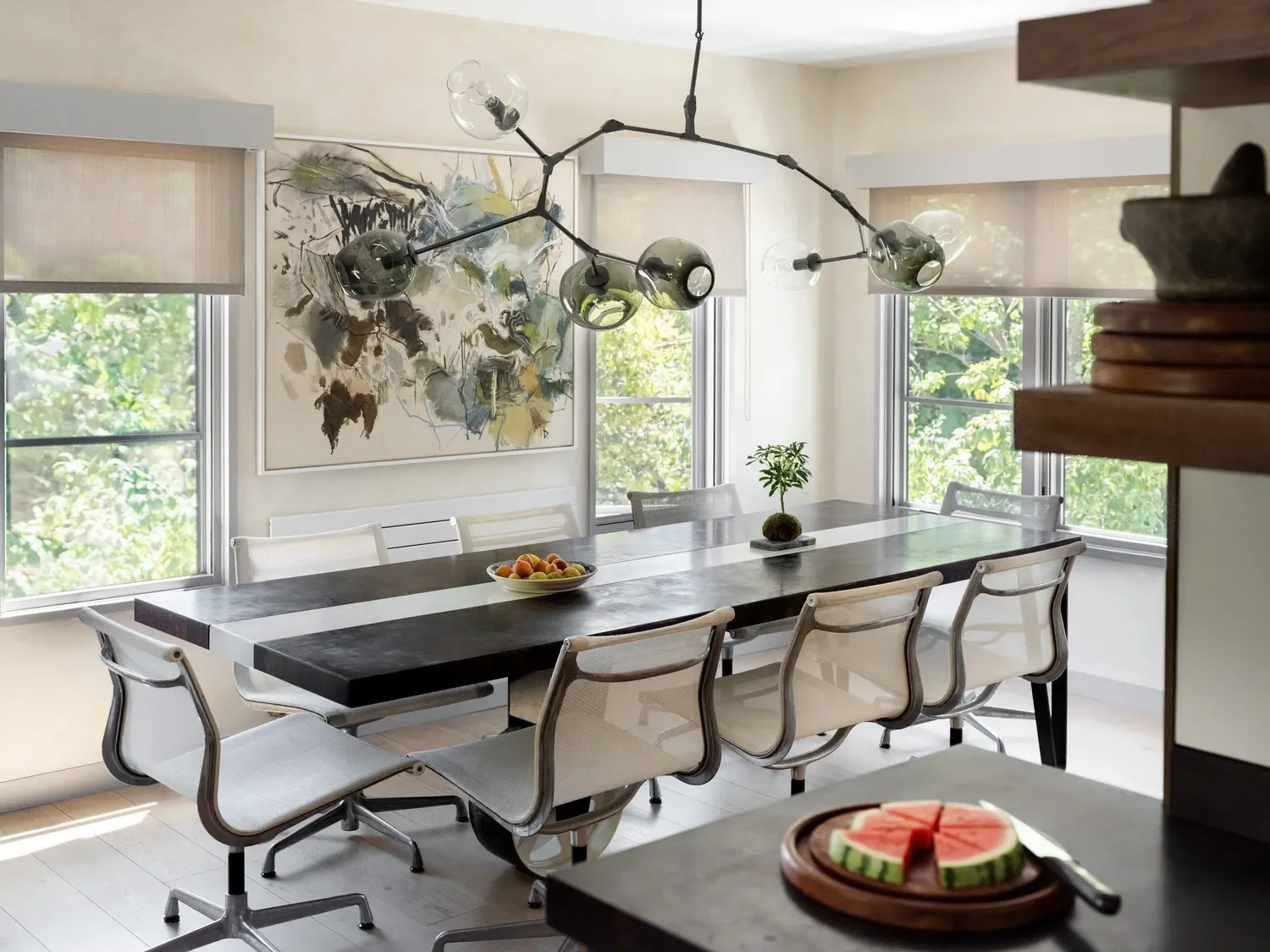 Modern dining room with a dark wood table, white chairs, large windows, a colorful abstract wall art, a contemporary chandelier, a fruit bowl, and a small potted plant.