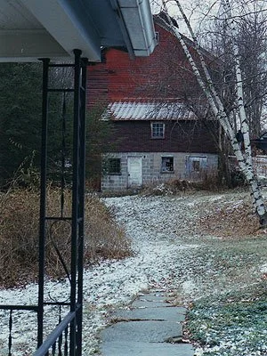 View of an old brick house with a metal roof, partially obscured by leafless trees, seen from a porch with a hanging chain, in a lightly snow-covered yard.