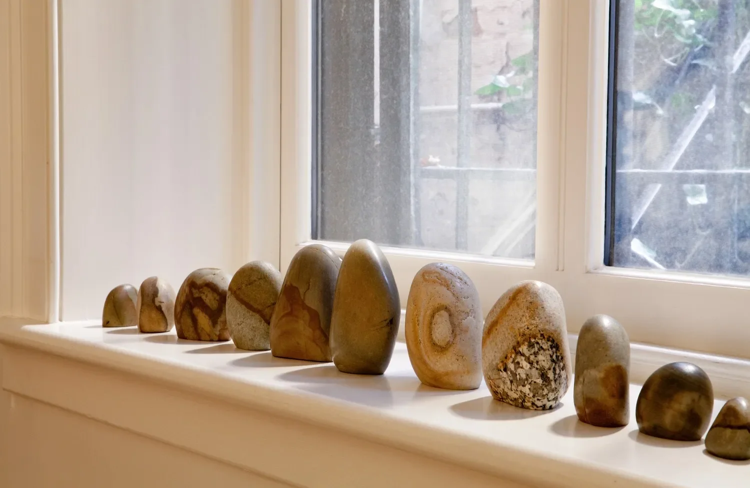 Collection of various small and medium-sized stones on a windowsill near a window with a view of a stone wall and outside plants.