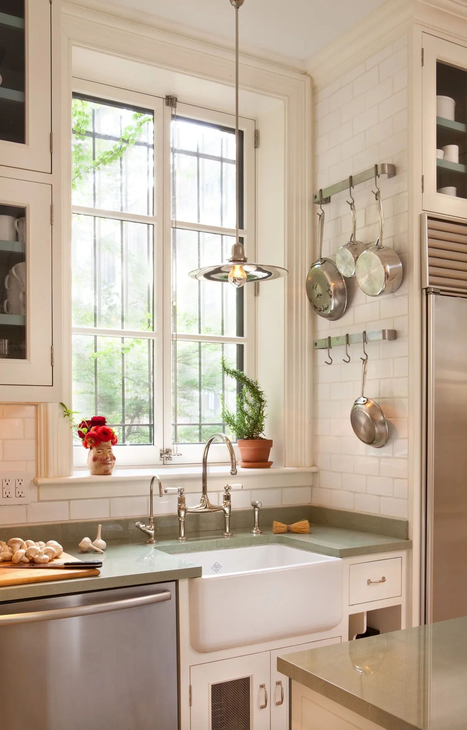 Kitchen scene with a large window, a white farmhouse sink, and greenery outside. Cookbook with garlic and a knife on the counter, decorative vase with red flowers, potted plant, hanging pots, and a vintage-style pendant light.