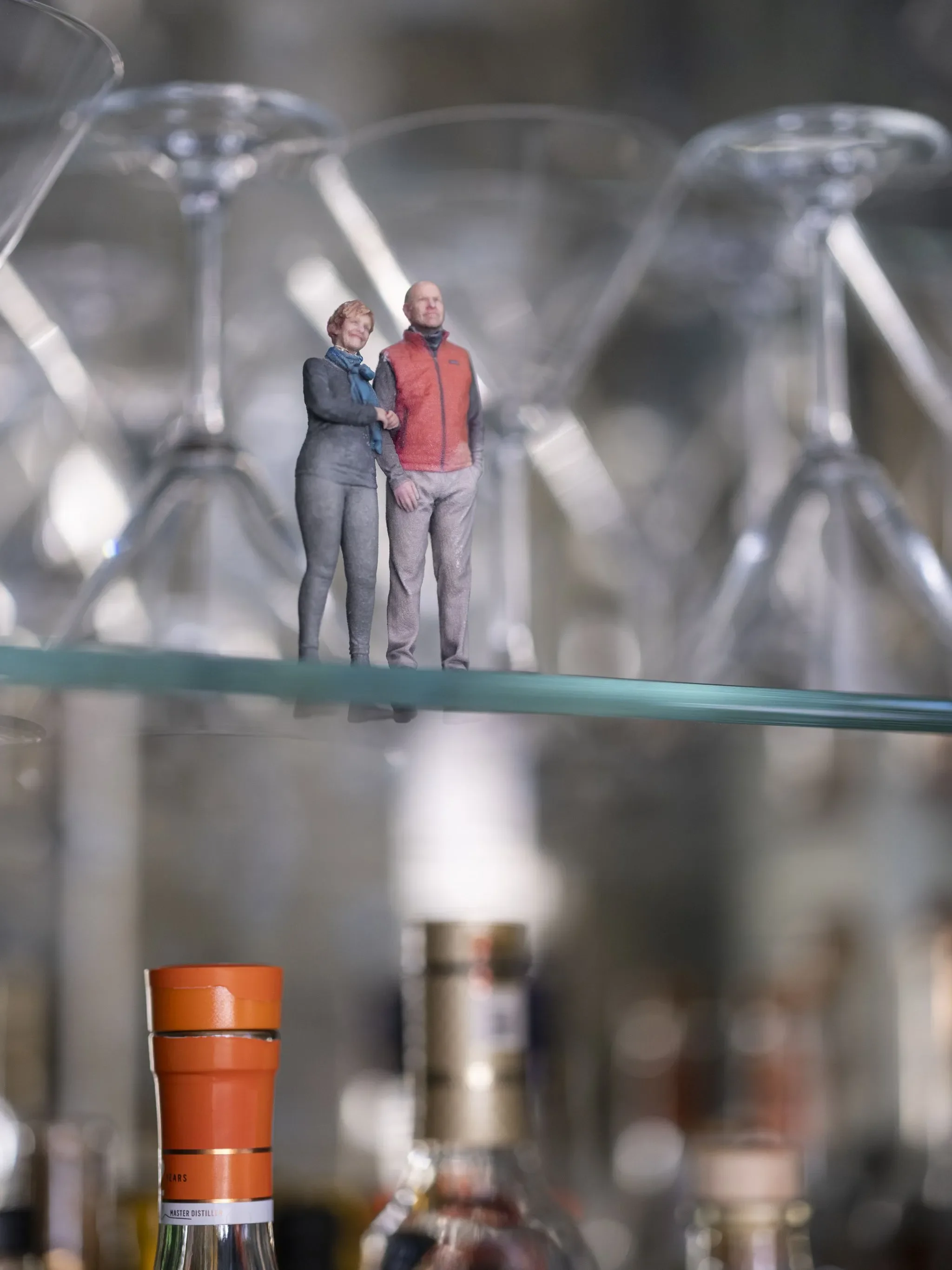 Miniature figurines of a man and woman shaking hands, standing among various bottles and glasses on a shelf.