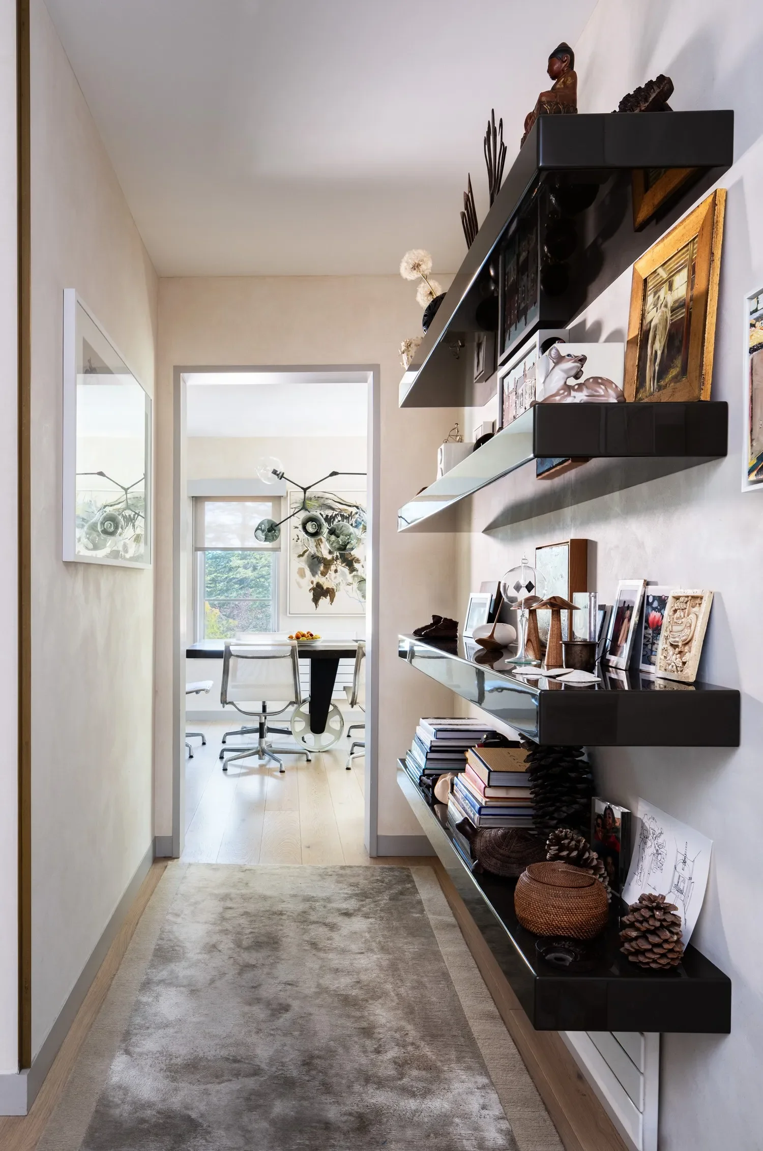 A cozy, well-decorated hallway leading to a dining area with a window, white walls, a beige rug, wall-mounted black shelves holding decorative items, framed artwork, and printed photographs.