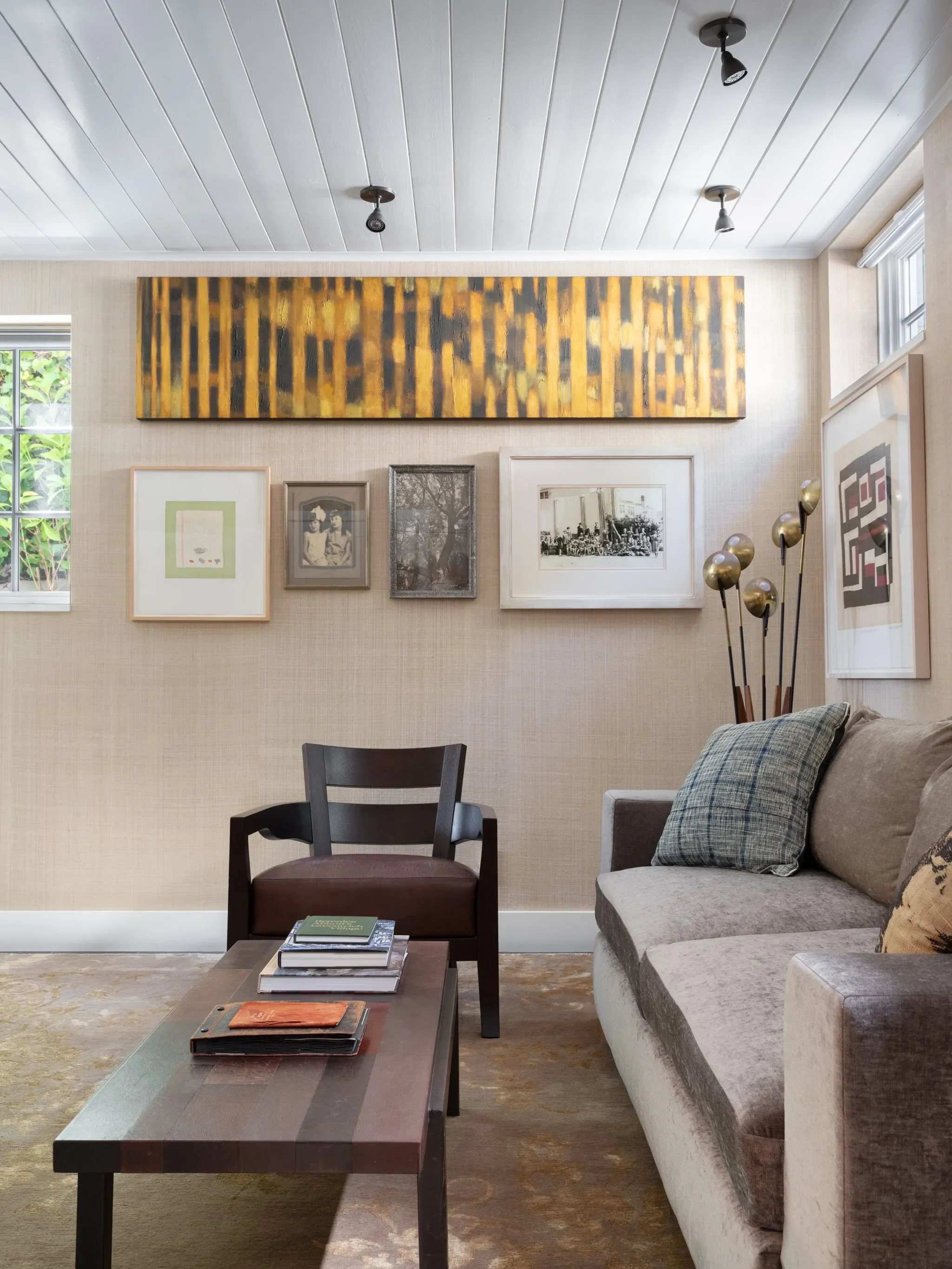 Living room with off-white wallpaper, framed photos and artwork on the wall, a gray couch with pillows, a black chair, and a wooden coffee table with books and a notebook. Large windows with white trim and a beige curtain. Stylish floor lamp with globe shades.