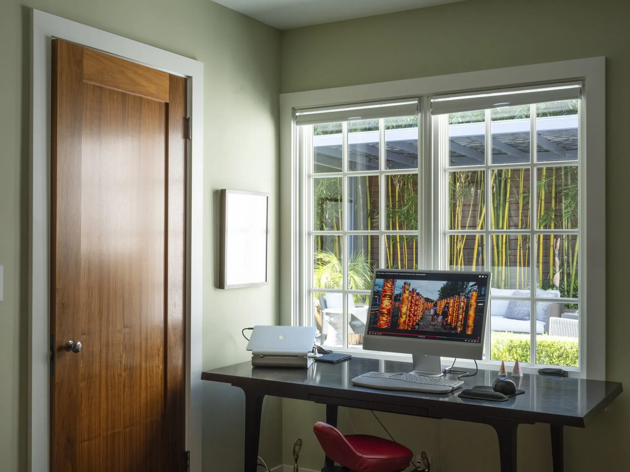 Home office space with a desk, an iMac computer displaying a cityscape at night, a red chair, a window with blinds showing greenery outside, and a closed wooden door.