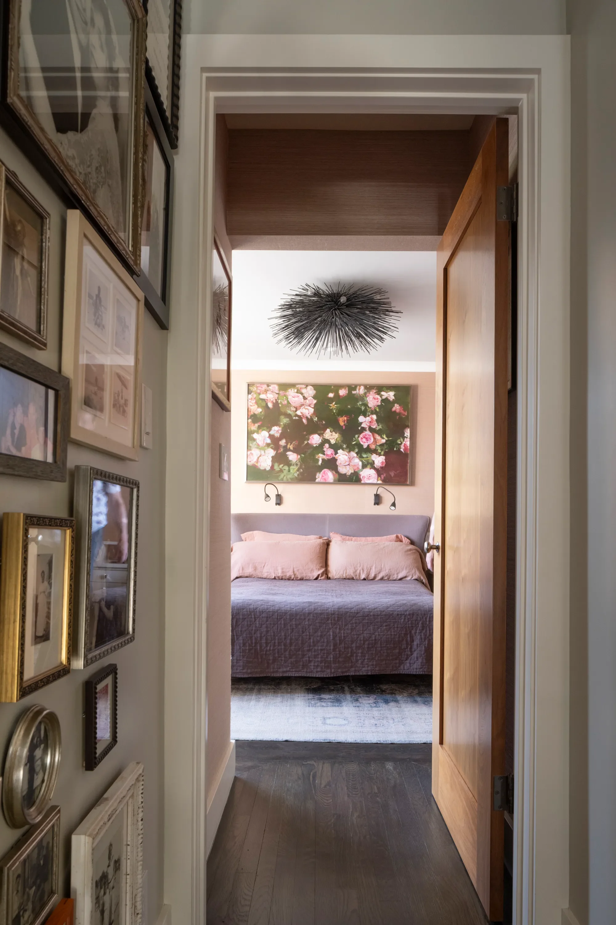 View through a doorway into a bedroom with pink bedding, floral painting above the bed, and a modern chandelier on the ceiling.