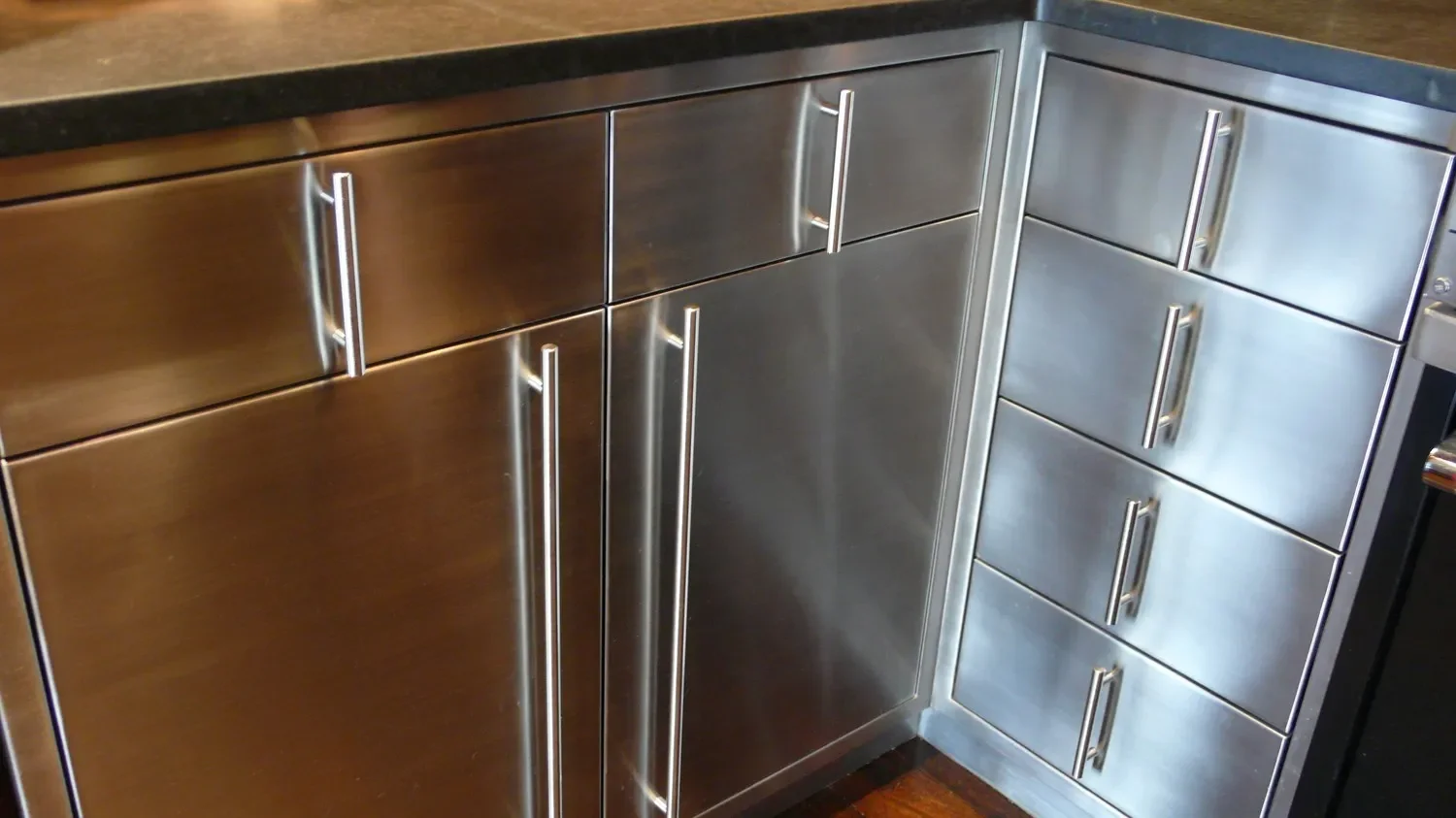 Stainless steel kitchen cabinets with long vertical handles, next to a dark countertop, in a modern kitchen setting.