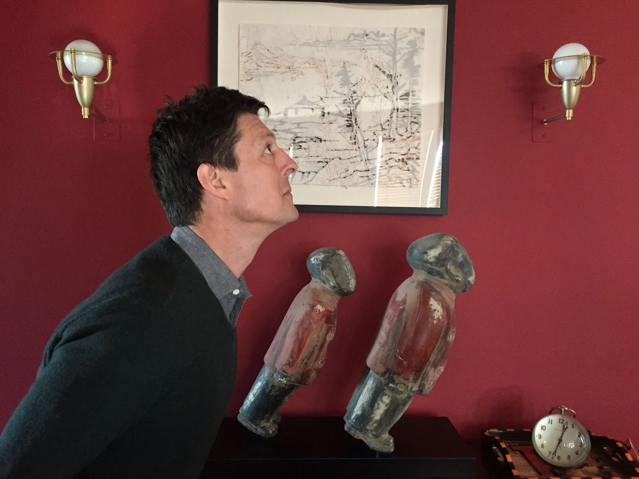 A man with dark hair, wearing a black sweater over a gray collared shirt, is looking upward in a room with red walls. Behind him are two abstract wooden sculptures shaped like elongated heads, a framed black-and-white landscape drawing, two wall-mounted white globe lights with brass fixtures, and a round clock on a table with a striped runner.
