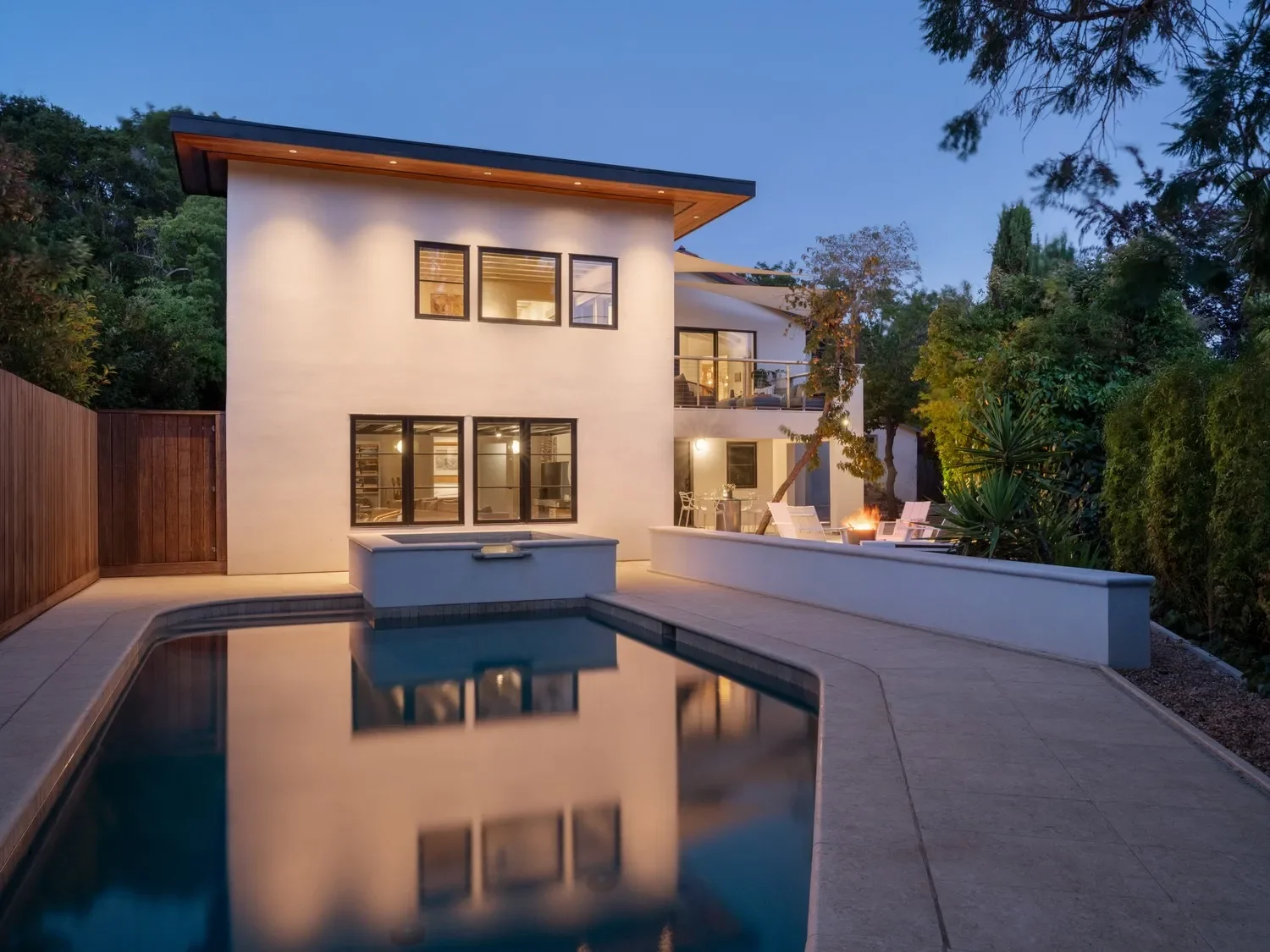 Modern two-story house with large windows and balcony, outdoor pool, and patio with seating and fire pit, surrounded by trees at dusk.