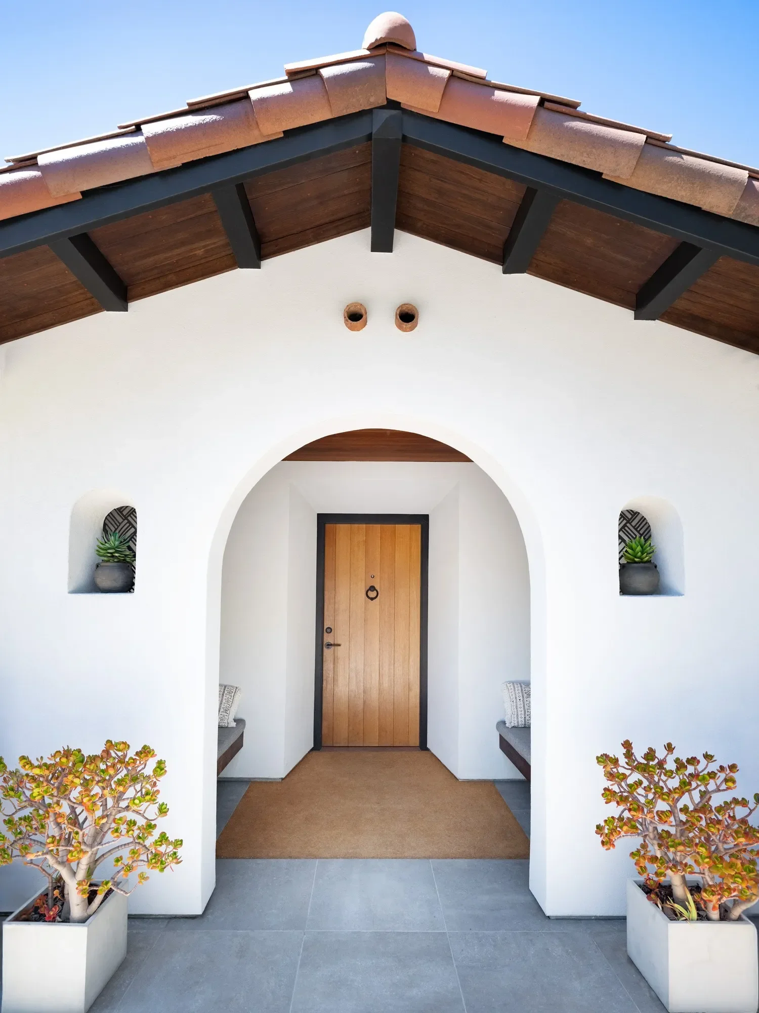 White stucco house entrance with a wooden door, arched alcoves with potted plants, and a covered porch with a tile roof. Decorative plants in square planters on each side of the entrance.