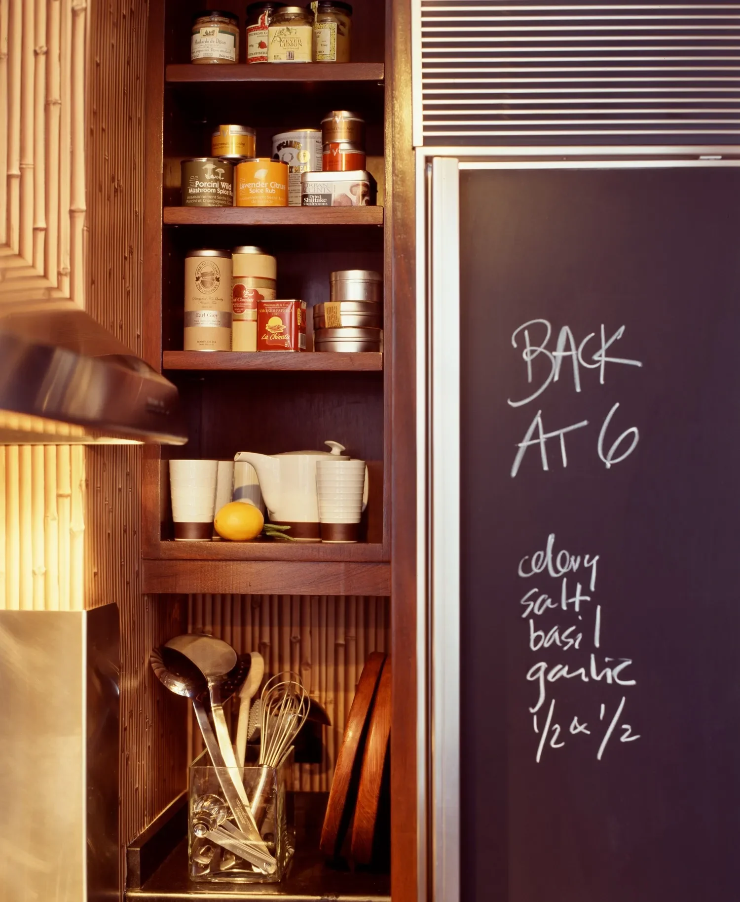 Kitchen cabinet shelf with jars of spices and herbs, cups, mugs, a lemon, and kitchen utensils, next to a chalkboard with a recipe written on it.