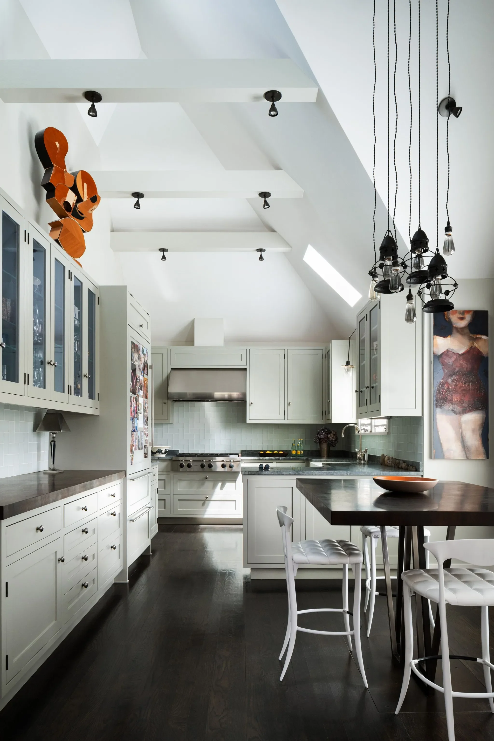A modern kitchen with white cabinets, dark wood floors, a black chandelier with exposed light bulbs, and a dining area with white chairs and a wooden table. There's artwork on the wall and a skylight letting in natural light.