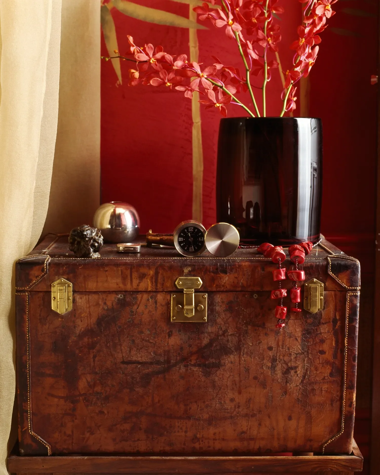 A vintage wooden chest with gold hardware, topped with decorative items including a black vase with pink orchids, a small silver object, a black clock, a pop of red beads, and a bronze sculpture, against a background of red and beige walls.