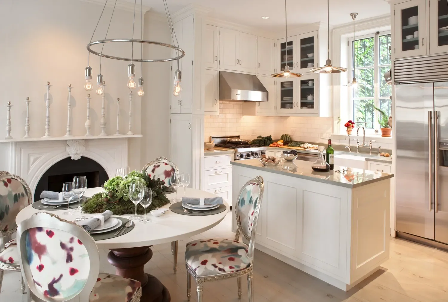 A bright, white kitchen and dining area with a round table set for a meal, surrounded by four upholstered chairs with colorful, abstract patterns. The table has wine glasses, napkins, and a floral centerpiece. The kitchen features white cabinetry, a kitchen island with a countertop, and a window letting in natural light, with some vegetables and kitchen items on the counter. Pendant lights hang above the island and dining table, and a stainless steel refrigerator is visible on the right.