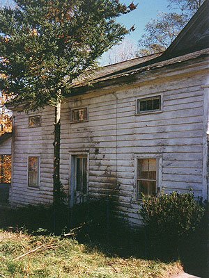 Old, weathered white wooden house with peeling paint, four small windows, and overgrown bushes in front.