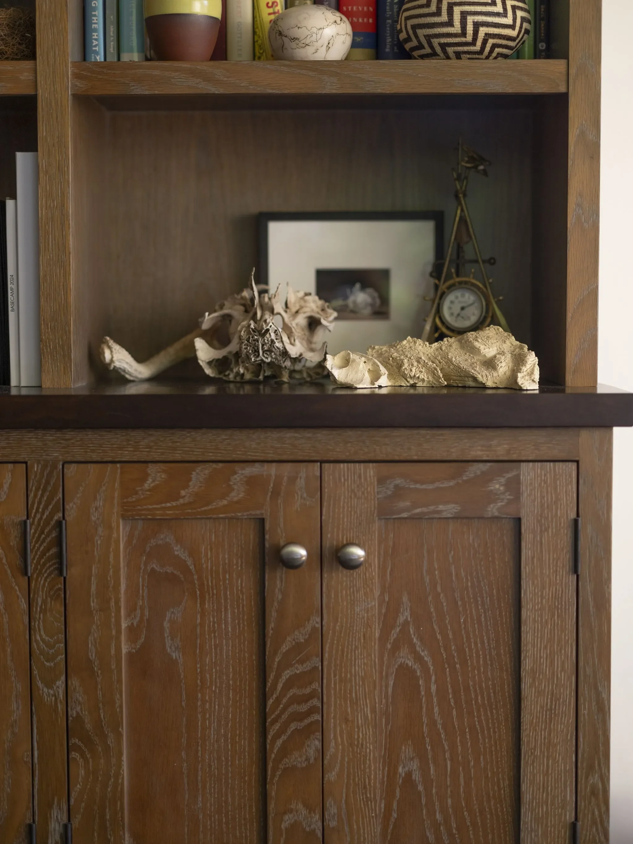 A wooden cabinet with shelves displaying books, vases, and decorative objects, including shells and a framed photo or artwork. There is a polished wooden surface with some seashells and a seashell fragment on top.