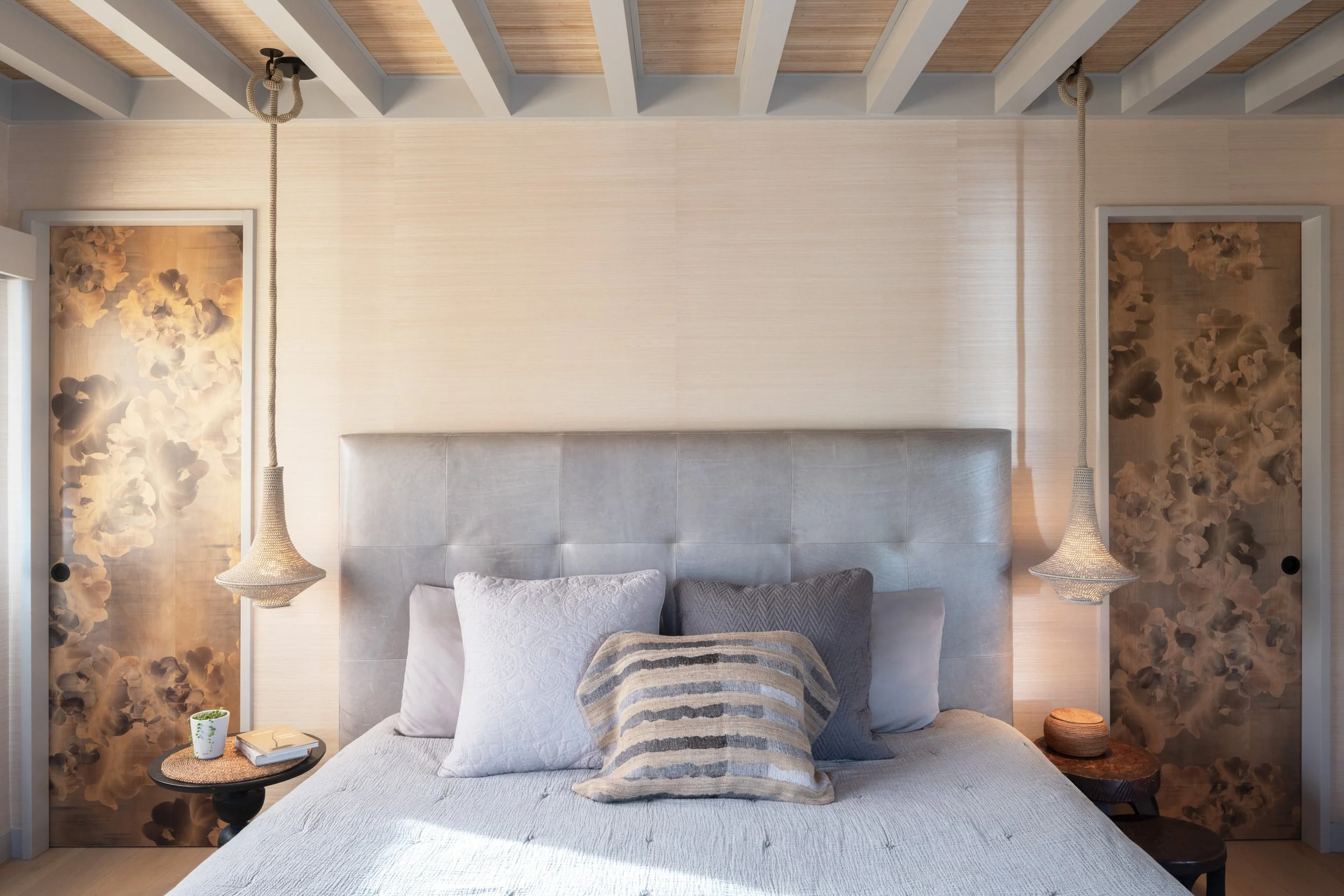 A bedroom with a large padded headboard, decorative pillows, hanging pendant lights, floral-printed sliding closet doors, a small side table with a plant, books, and a round decorative item.
