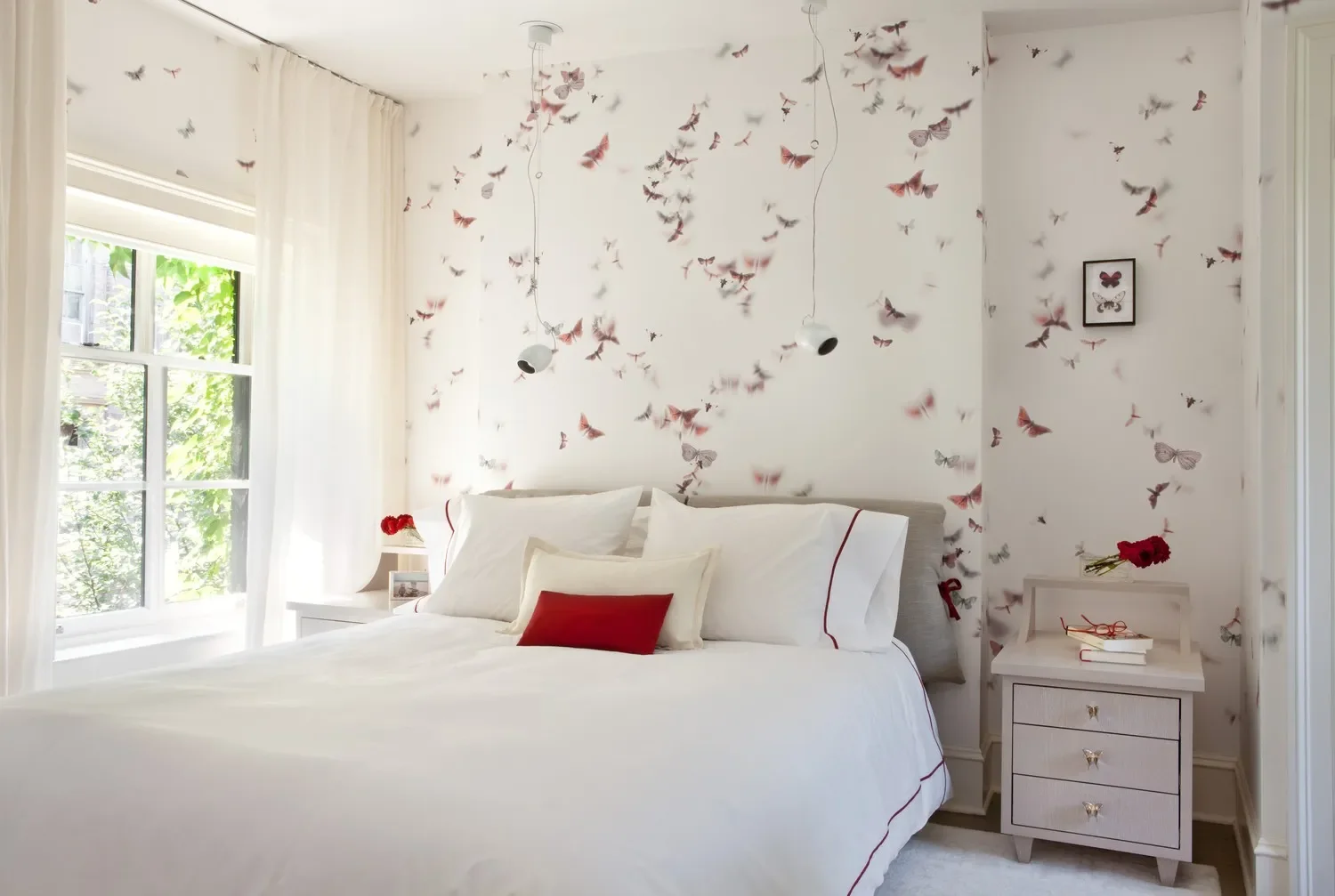 Bedroom with white bedding and pillows, butterfly-themed wallpaper, two hanging pendant lights, nightstand with red flowers and books, and a framed butterfly picture on the wall.