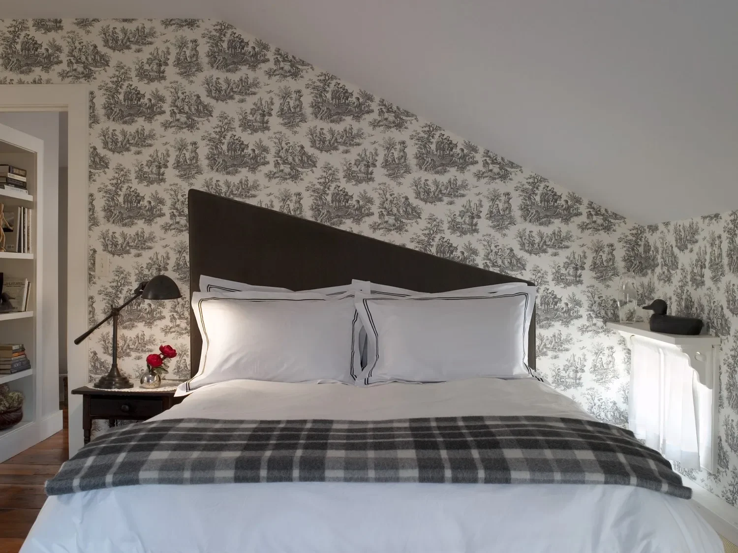 A bedroom with a sloped ceiling, featuring a bed with white pillows and a checkered blanket. The wall behind the bed has black and white toile wallpaper. On the left side, a small wooden nightstand with a black lamp and a vase with red flowers. To the right, a white shelf with a black duck figurine and glass container, and a white radiator beneath the shelf.