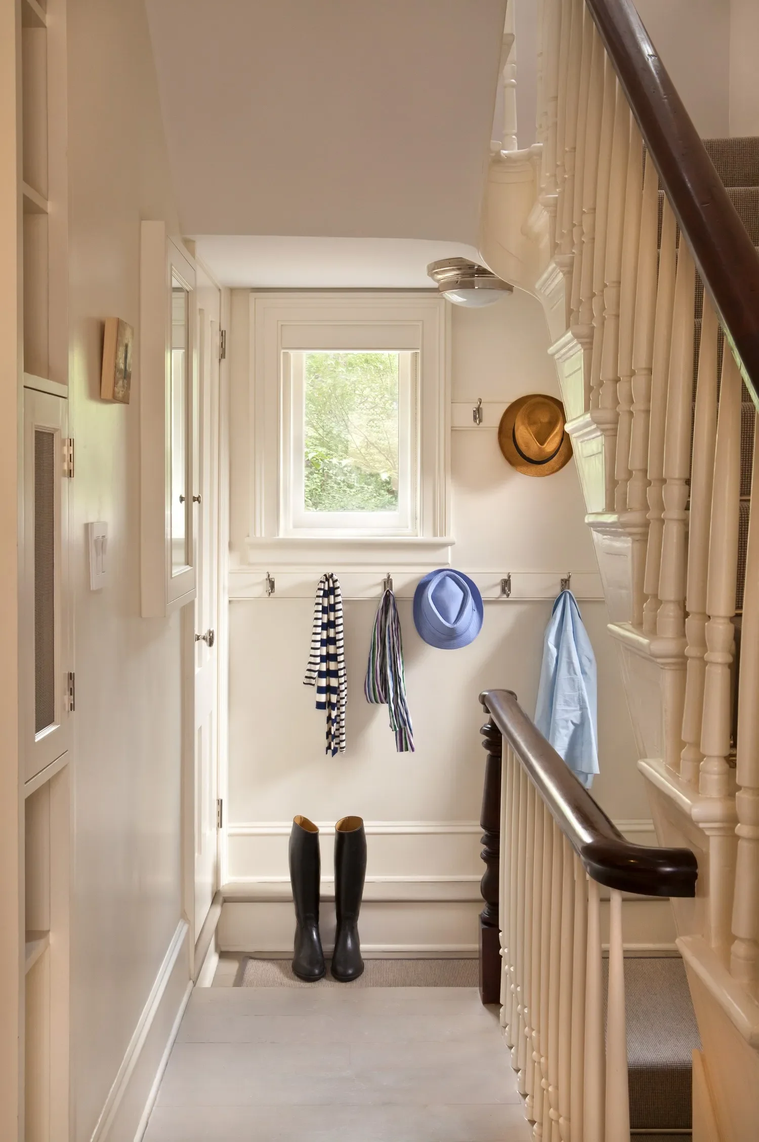 A hallway with a window, white walls, and a staircase on the right. Shoes, including black rain boots, are at the bottom of the stairs. Several hooks with hats and scarves are on the wall, and a wooden railing runs along the staircase.