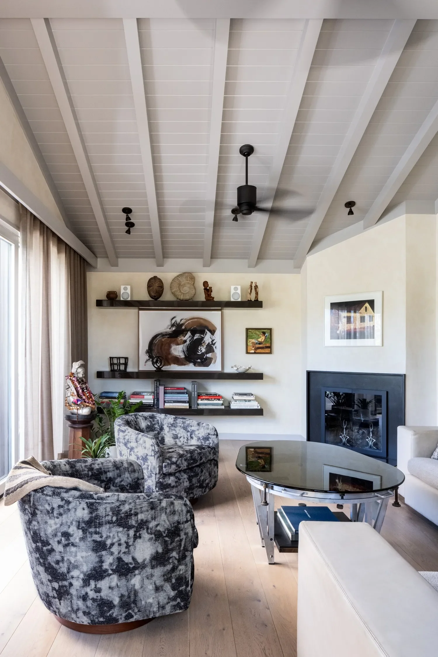 Living room with gray patterned armchair, glass coffee table, white couch, wall-mounted black shelves with decor and books, artwork, and ceiling fans.