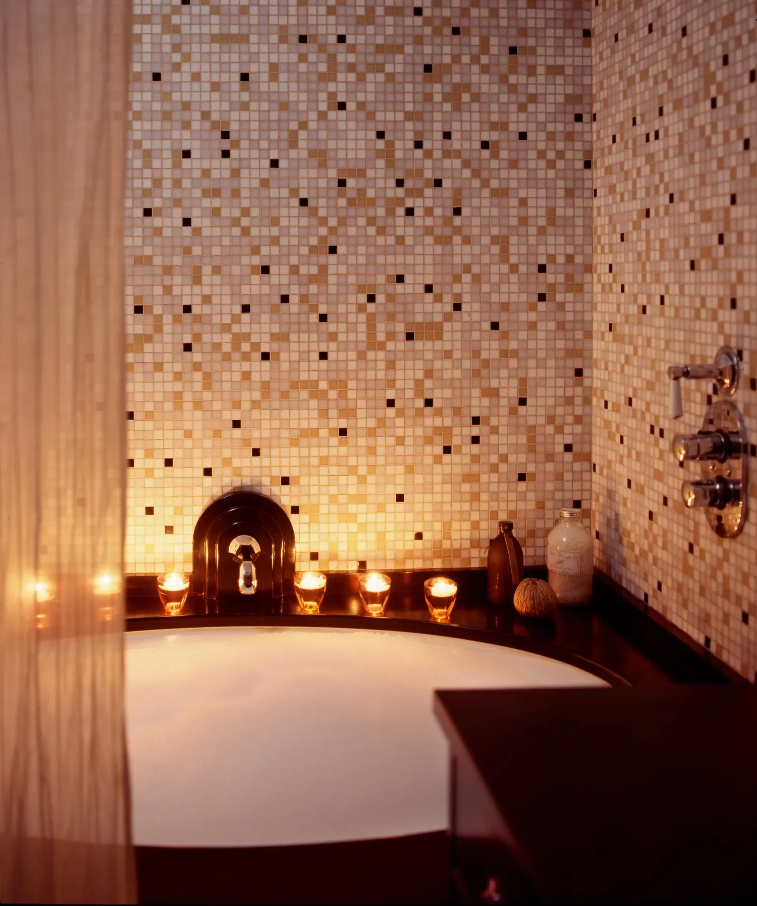 A bathroom with a tiled wall, candles surrounding a bathtub, and various bottles and natural items on the dark countertop.