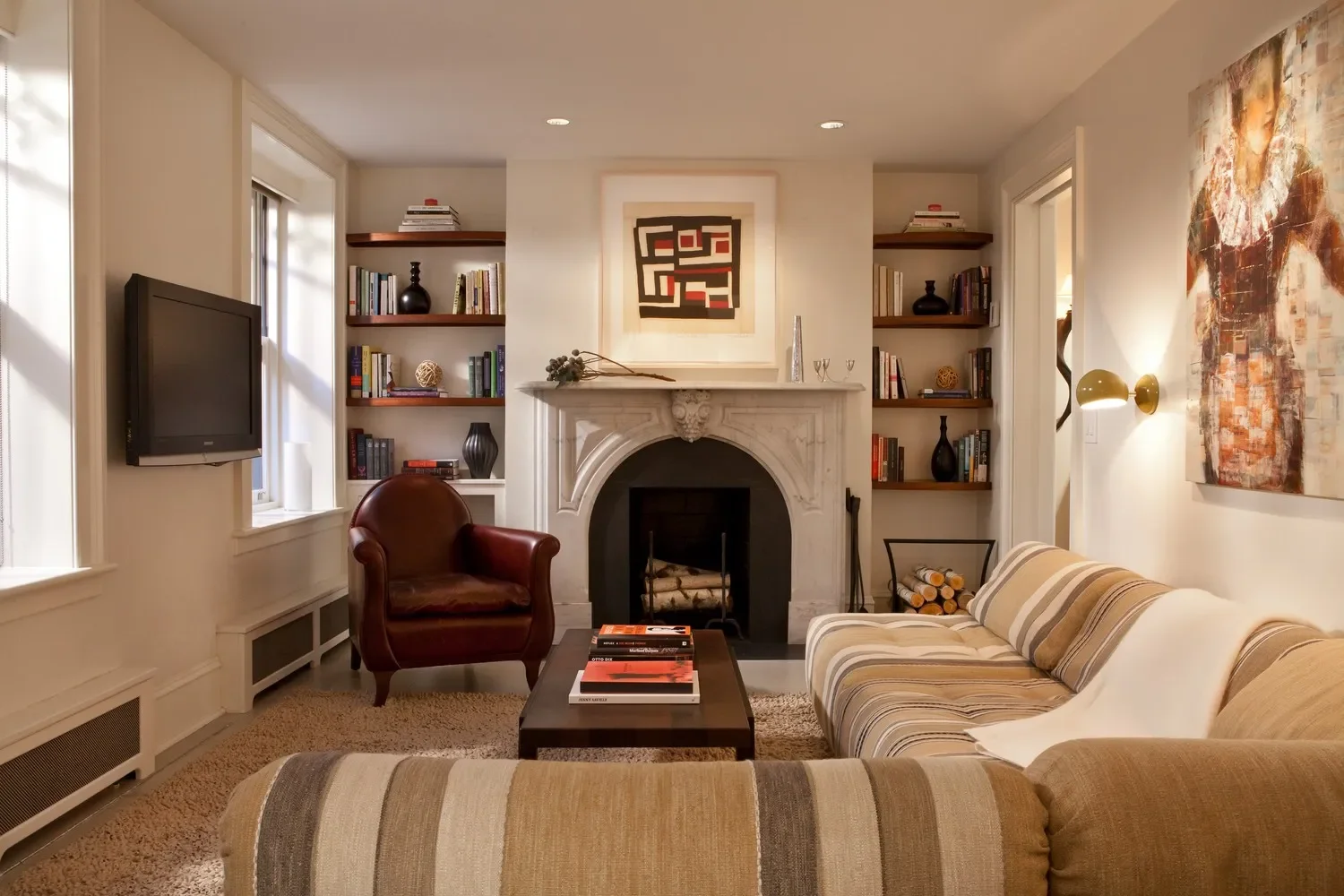 Living room with fireplace, bookshelves, striped sofa, leather armchair, wall art, wall-mounted TV, coffee table, and decorative items.