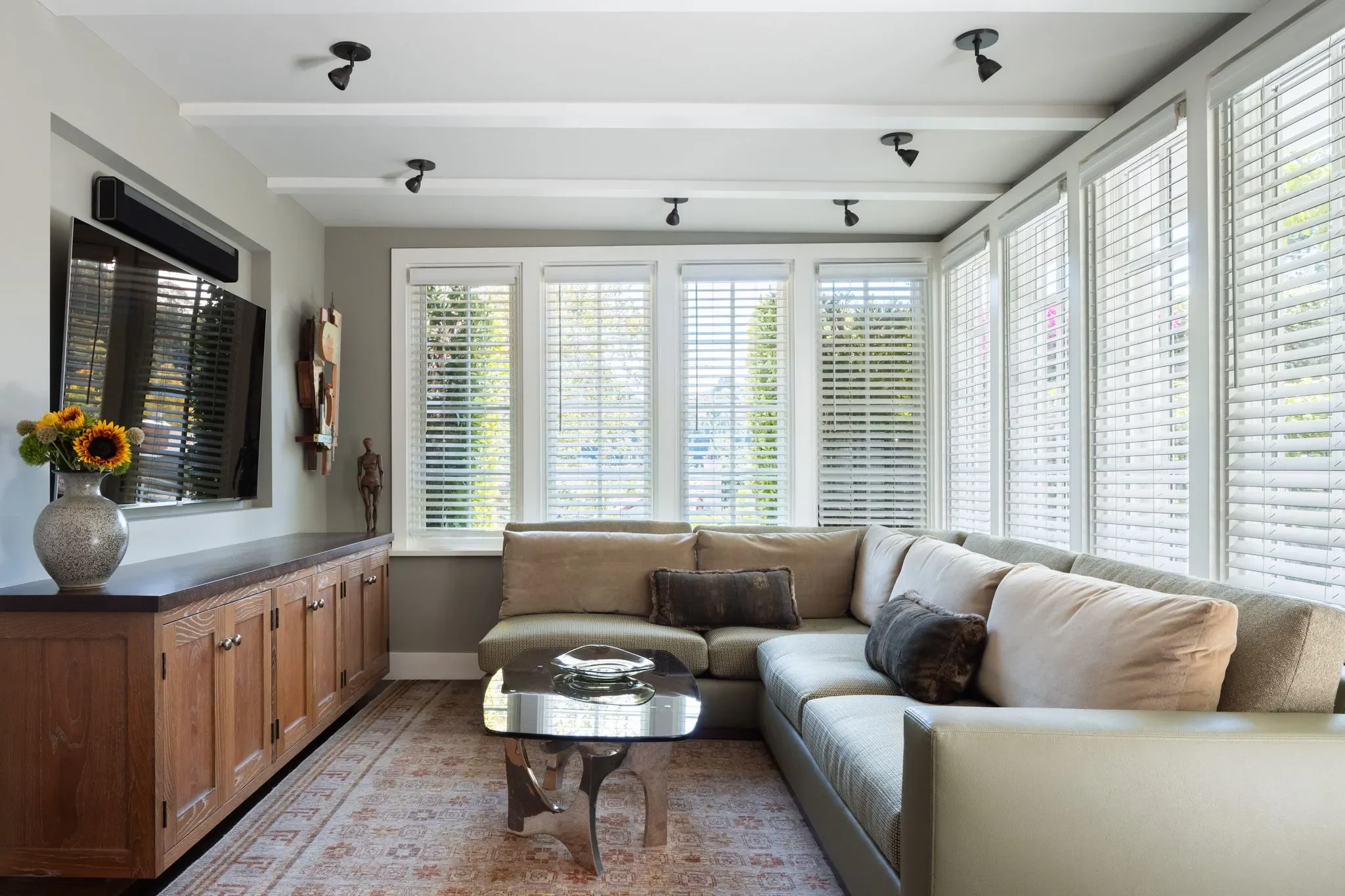 Living room with a beige sectional sofa, a coffee table, large windows with white blinds, a flat-screen TV on the wall, a vase with sunflowers, and a wooden cabinet.
