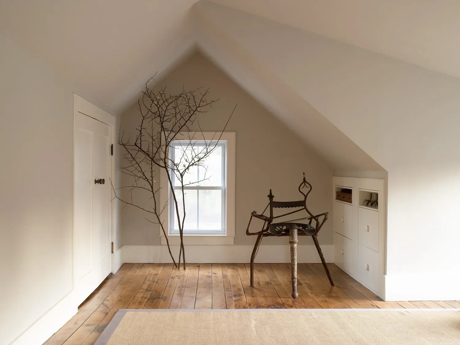 A corner of a room with a sloped ceiling, white walls, and hardwood floor. There is a window, a small leafless tree branch, an antique iron chair, and a shadowed built-in cabinet.