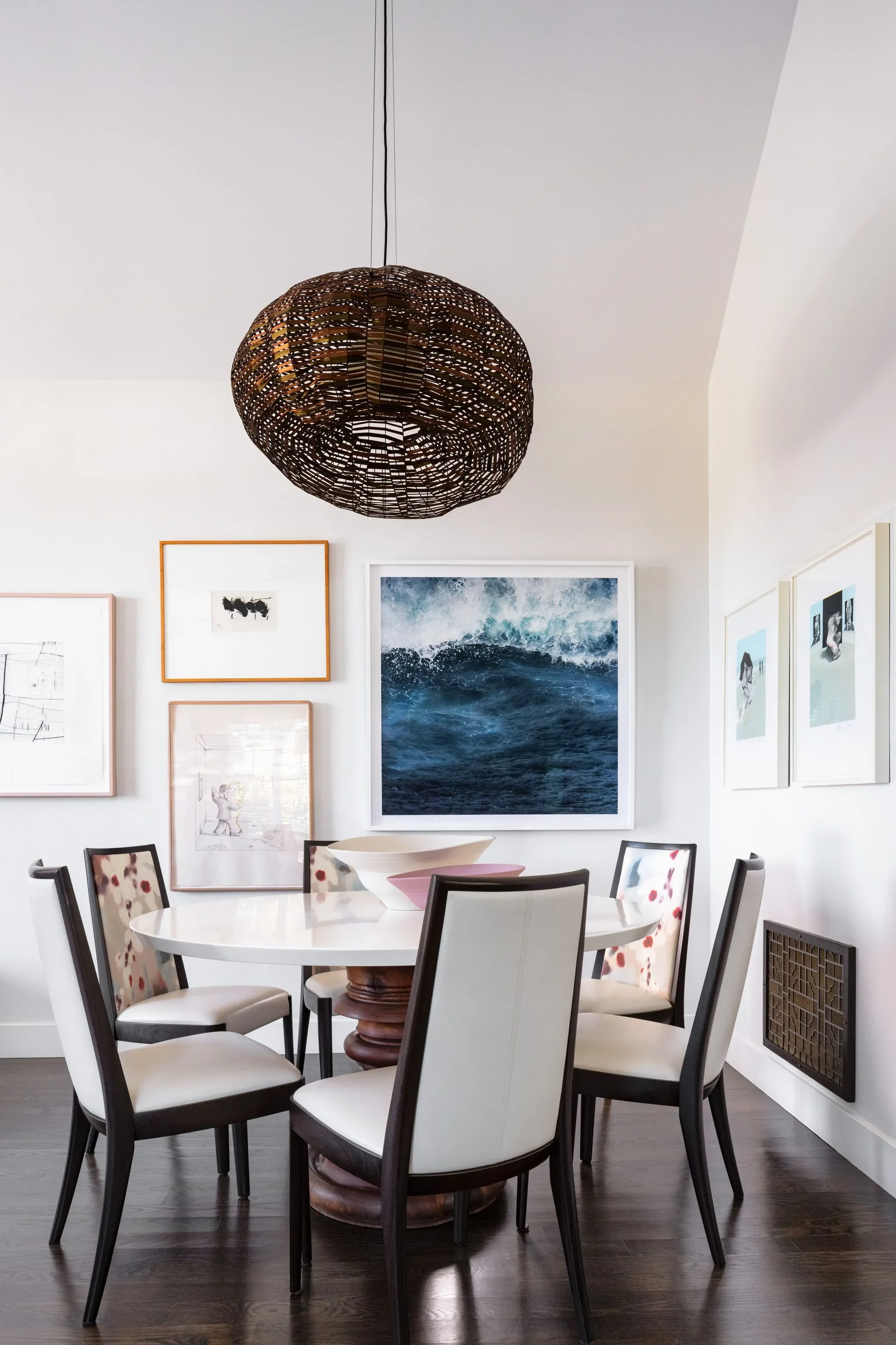 A round white dining table surrounded by eight chairs with black frames and floral patterned upholstery, in a room with framed artwork on white walls and a dark wooden floor. A woven pendant light hangs from the ceiling.