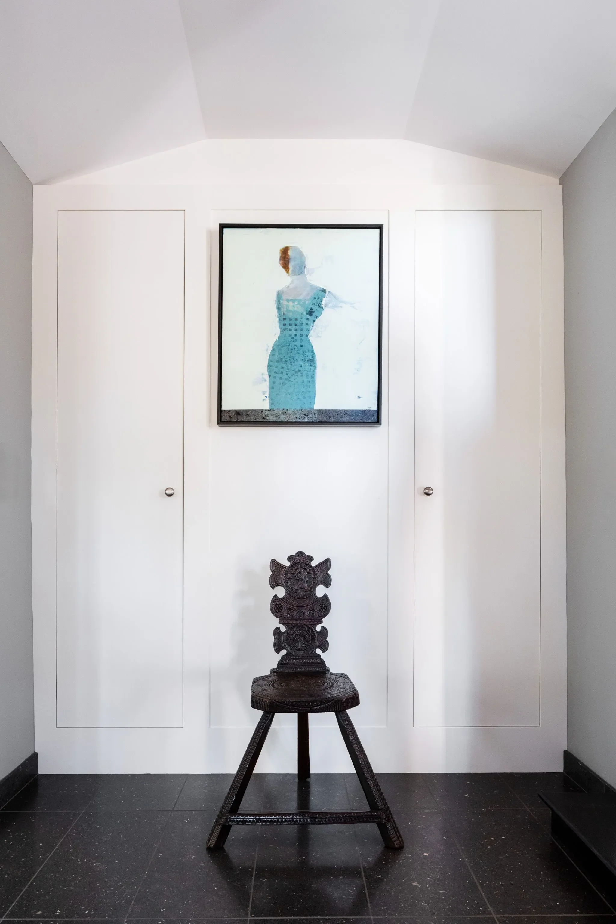 A room with white walls, a framed portrait of a woman in a blue dress hung on the wall, an antique carved wooden chair with a dark finish centered in front of the wall, and black tiled floor.