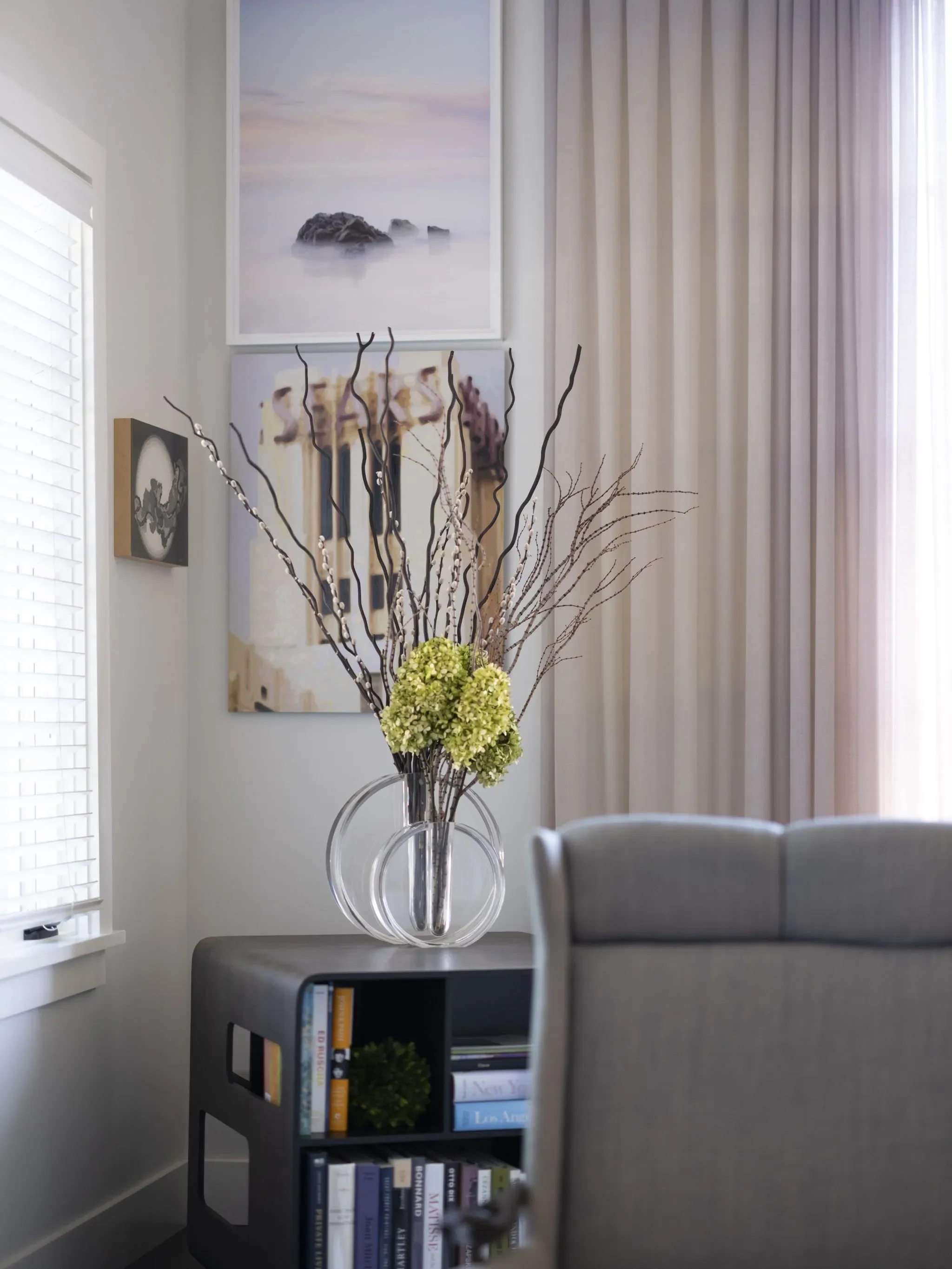 A living room corner with artwork on the wall, a glass vase with decorative branches and green hydrangea flowers on a table, a bookshelf, and beige curtains letting in natural light.