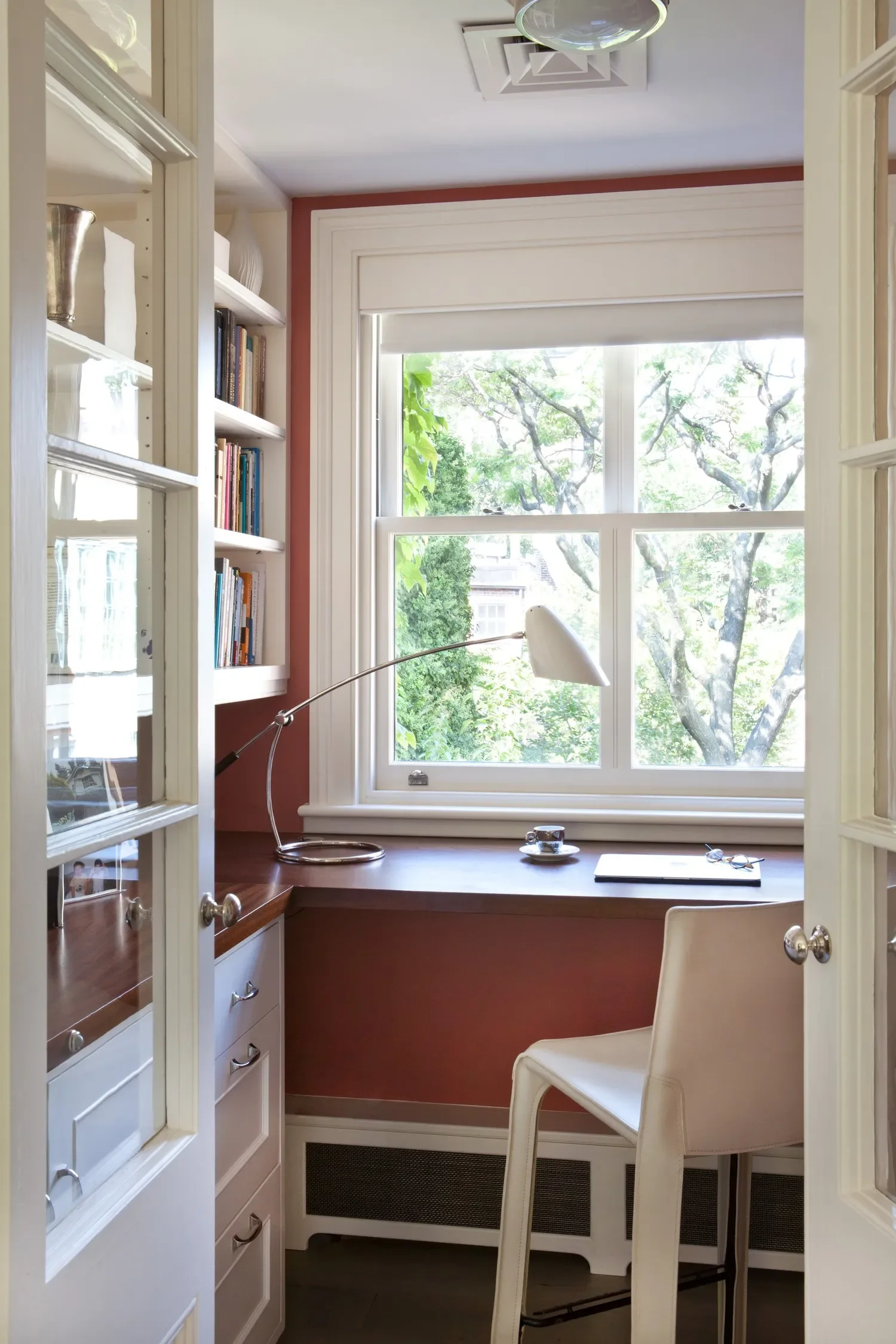 A cozy workspace with a large window overlooking green trees. There's a white chair, a wooden desk with a black notebook, eyeglasses, a cup, and a tall white floor lamp. On the left, a white bookshelf with books and decorative items, and a painting or framed picture. The room has white trim and a reddish-brown wall.