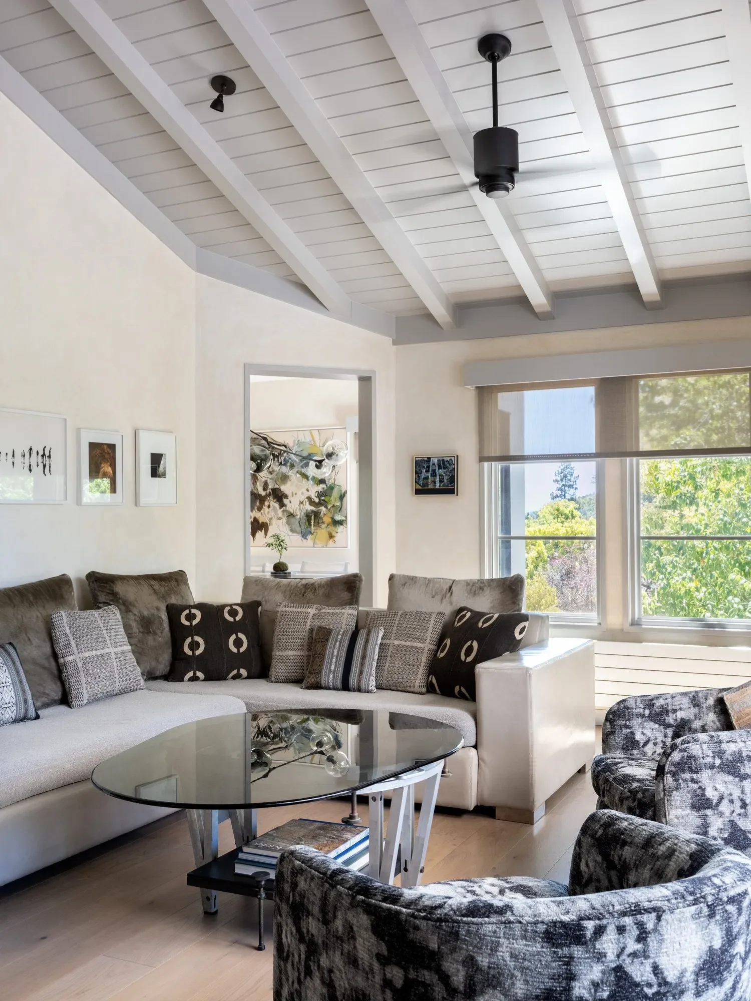 A living room with a white sectional sofa, various patterned pillows, a glass coffee table, and a black and gray armchair. Large windows with view of trees outside, white walls with framed art, and a white wooden ceiling with black ceiling fan and light fixture.