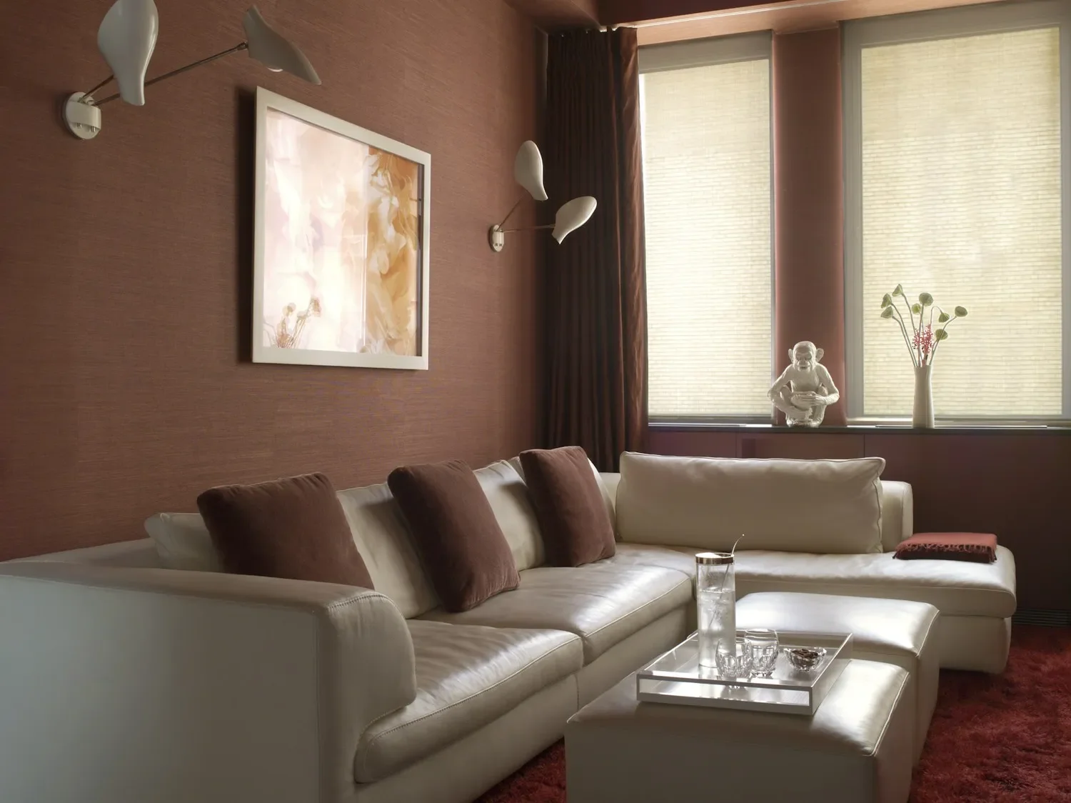 Living room with white sectional sofa, brown pillows, glass water pitcher, decorative glassware, and a tray on a white coffee table. The wall behind has a framed artwork and wall-mounted white reading lamps. Large windows with beige blinds and dark curtains, decorated with a white sculpture and a vase with flowers on the window sill.