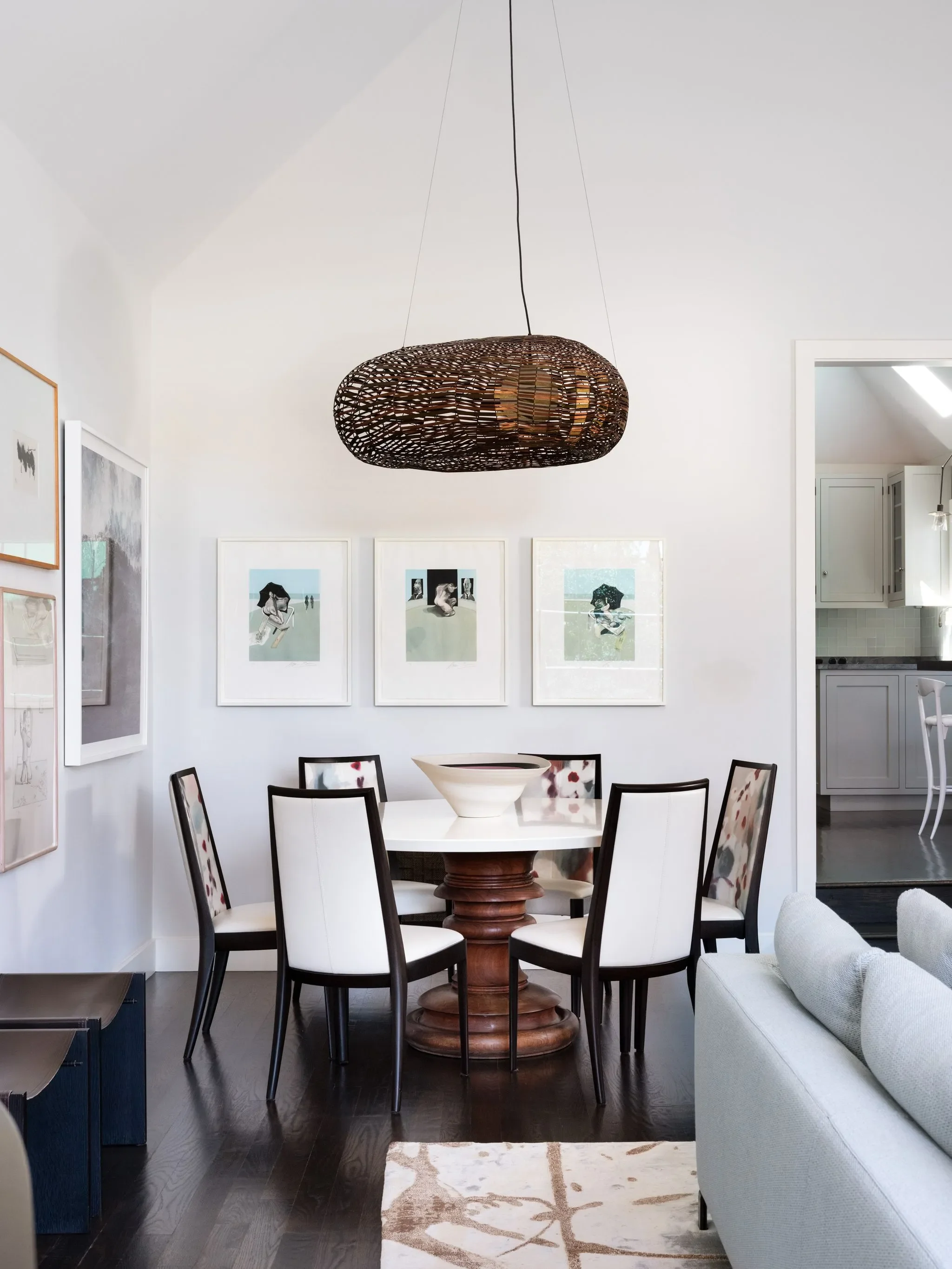 A dining room with a round table, eight chairs, framed artwork on white walls, a wicker ceiling light fixture, and a kitchen visible through an open doorway.