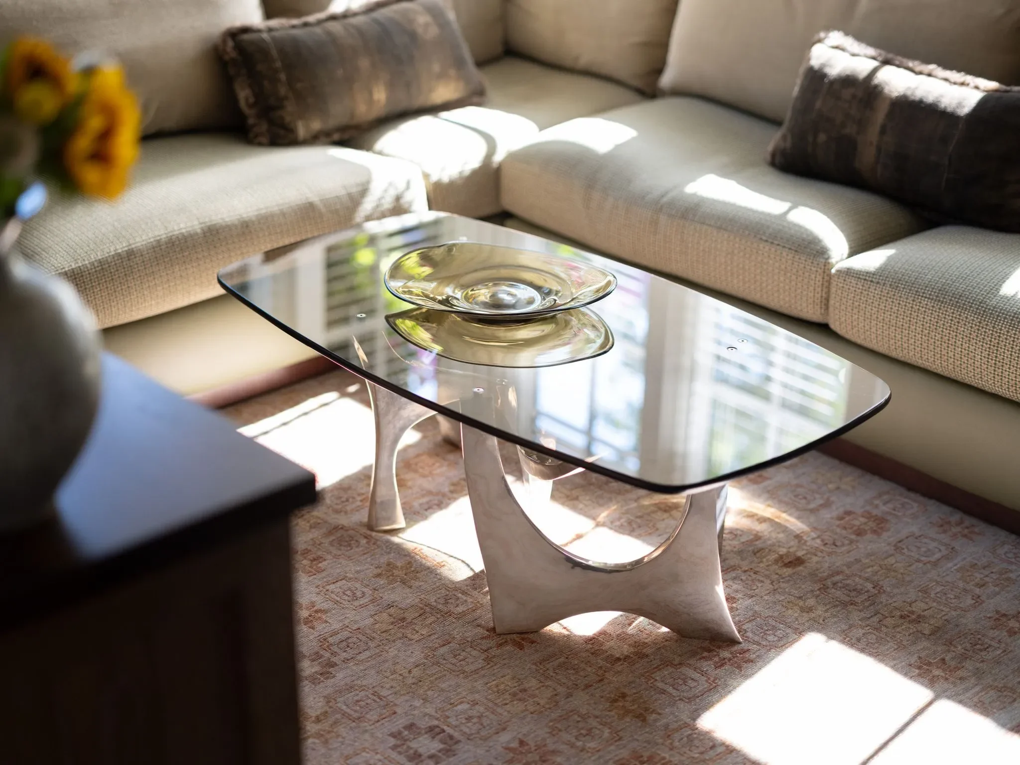 A living room with a beige sectional sofa decorated with dark patterned pillows. There is sunlight streaming through the window, casting shadows on the sofa. In front of the sofa, there is a glass coffee table with a sculptural, white base and a reflective surface.