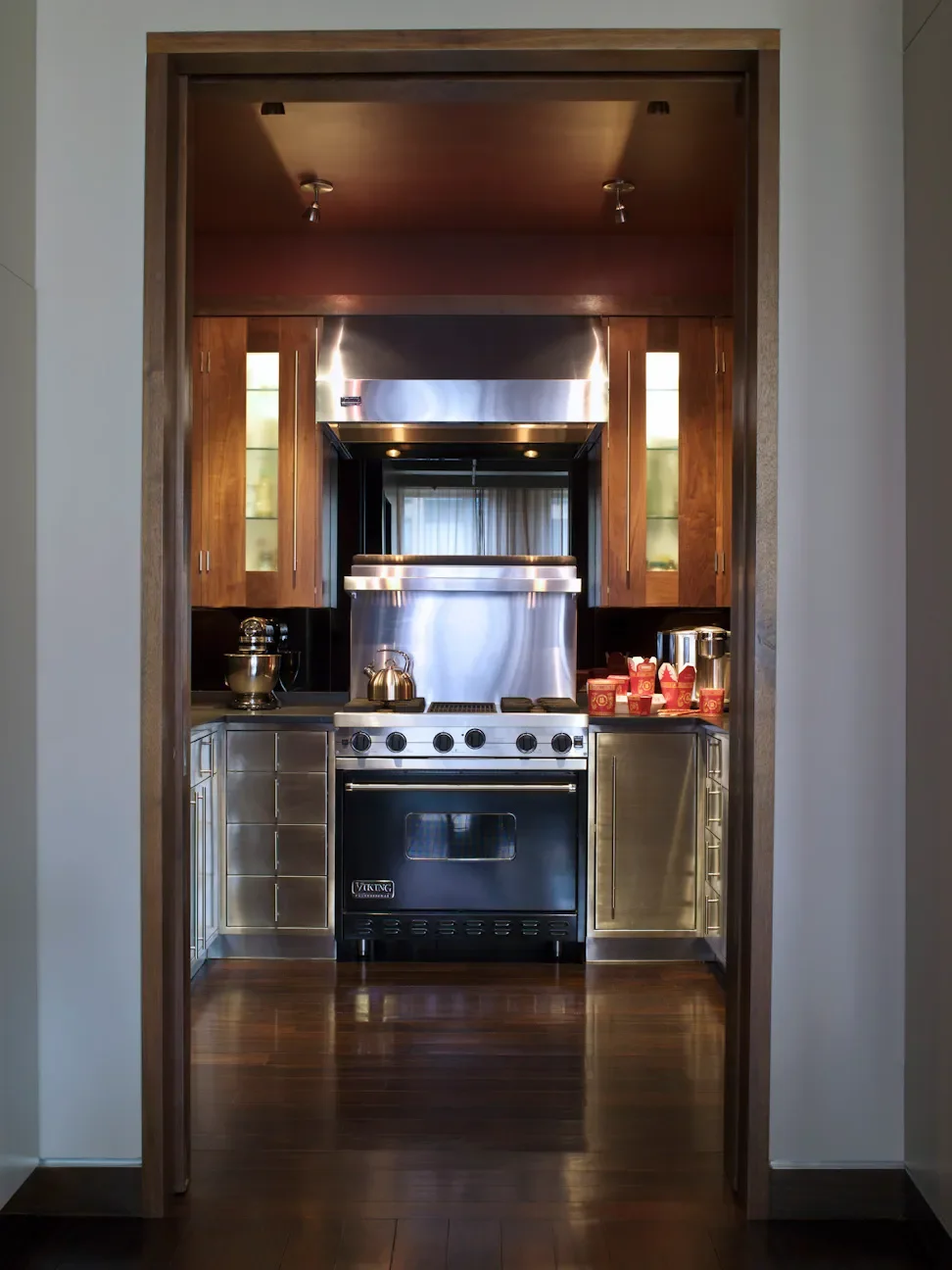 Kitchen view through a doorway with white walls and dark wood flooring, showing stainless steel appliances, wooden cabinets, and a glimpse of a dining area with curtains.