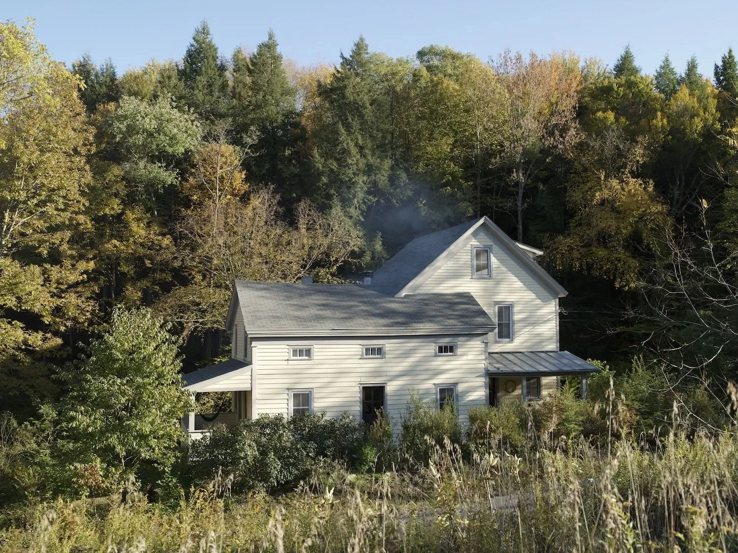 A two-story white house with gray roof nestled among trees on a hillside with autumn foliage, smoke rising from the chimney.