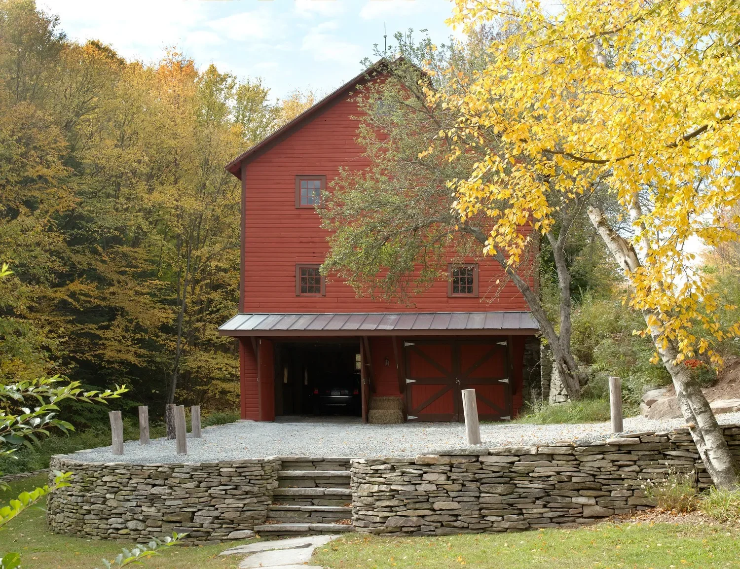 A red wooden barn with a gray metal roof, surrounded by autumn trees with yellow and green leaves, and a gravel driveway in the front.