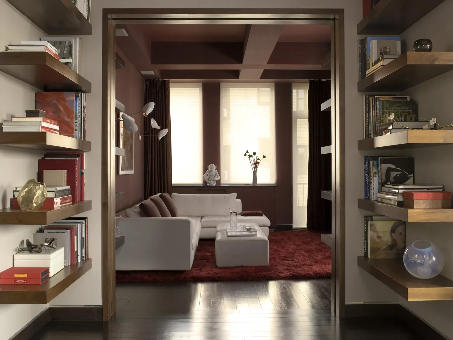 Living room viewed through doorway with built-in shelves on each side filled with books and decorative items. The room features a white sectional sofa, a red rug, and large windows with beige blinds, dark curtains, and a vase with flowers on the windowsill.