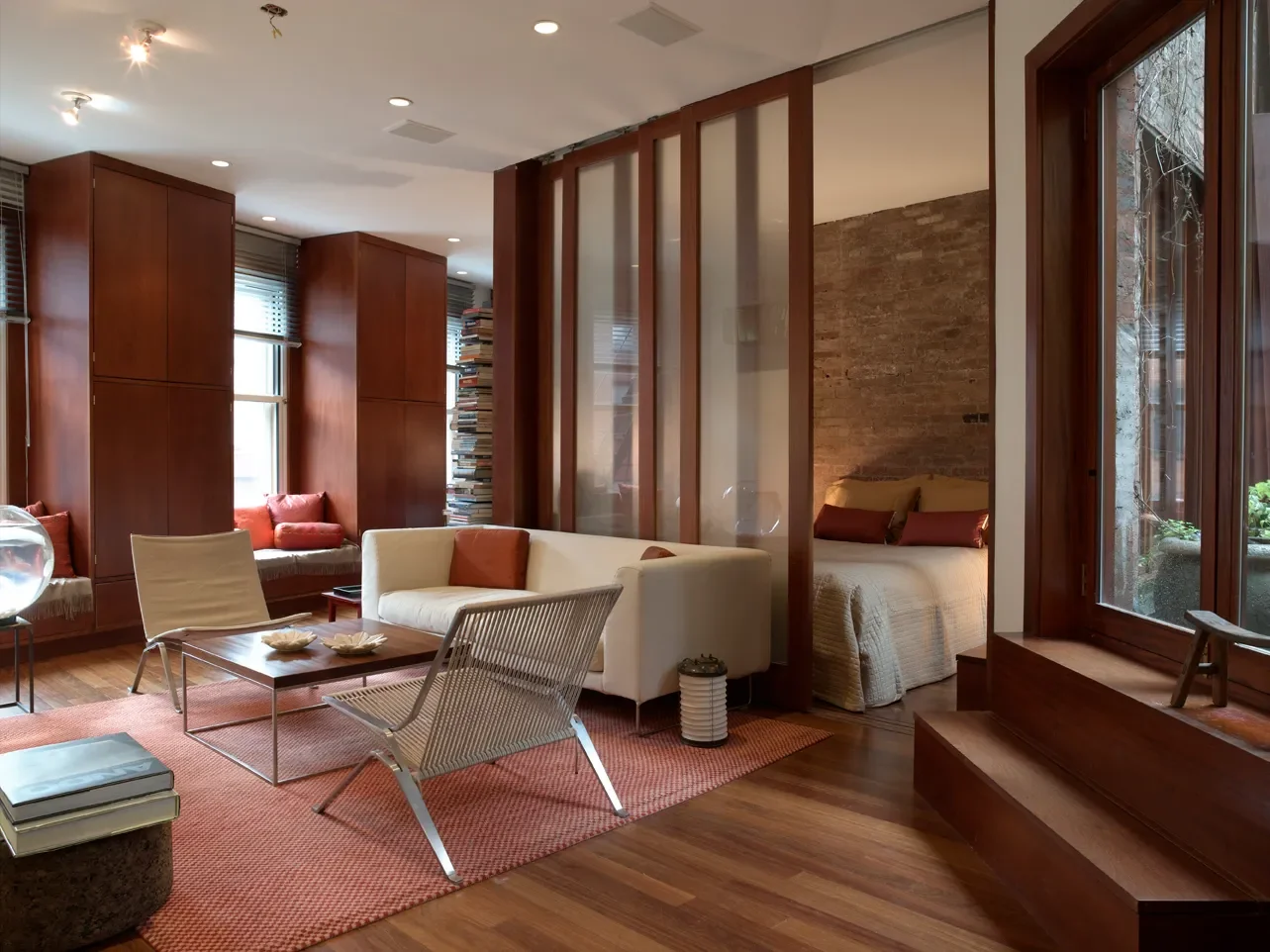 Living room with wooden floors, brick wall, white sofa, modern chairs, a table, built-in bookshelves, and a bed separated by wooden slats.