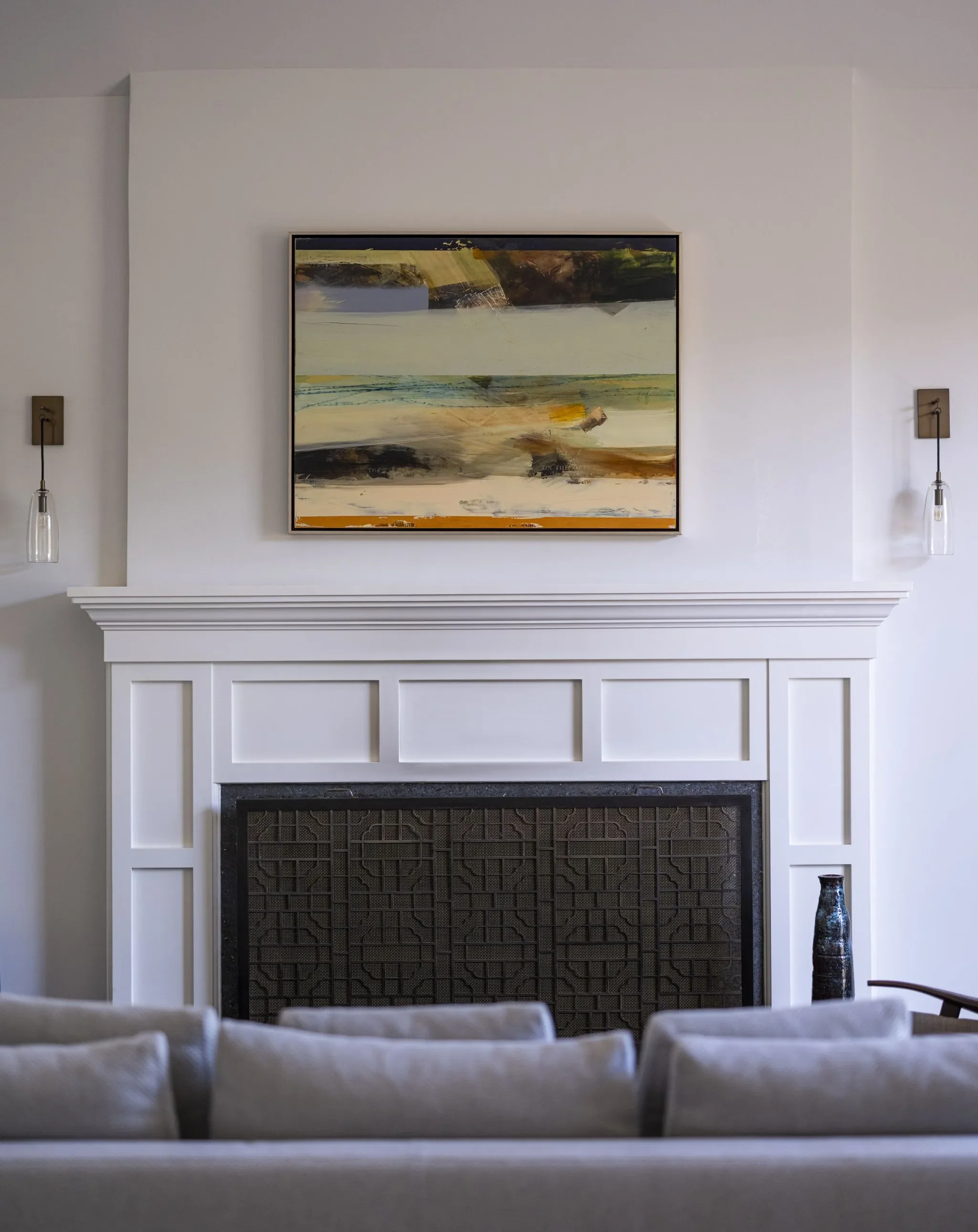 Living room with a white fireplace, abstract painting above it, two pendant lights on either side, and a blue vase on the right.