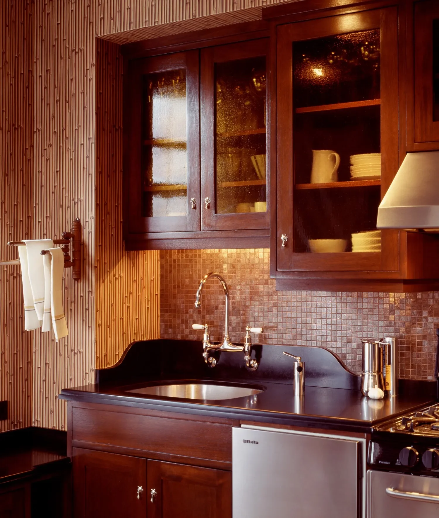 Kitchen with dark wood cabinets, a black countertop, a stainless steel sink, a chrome faucet, a tiled backsplash, and a wall with wood paneling. There are shelves with dishes behind glass cabinet doors, stainless steel containers on the counter, and a stove partially visible.