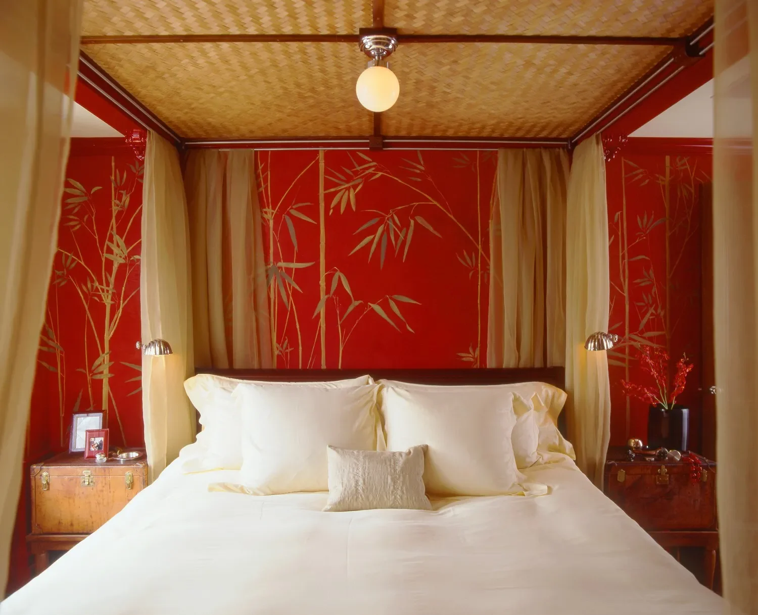 A bedroom with a white bed, four pillows, a red wallpaper with gold bamboo design, and beige curtains on both sides of the bed. There are two small bedside tables with lamps and decorative items.