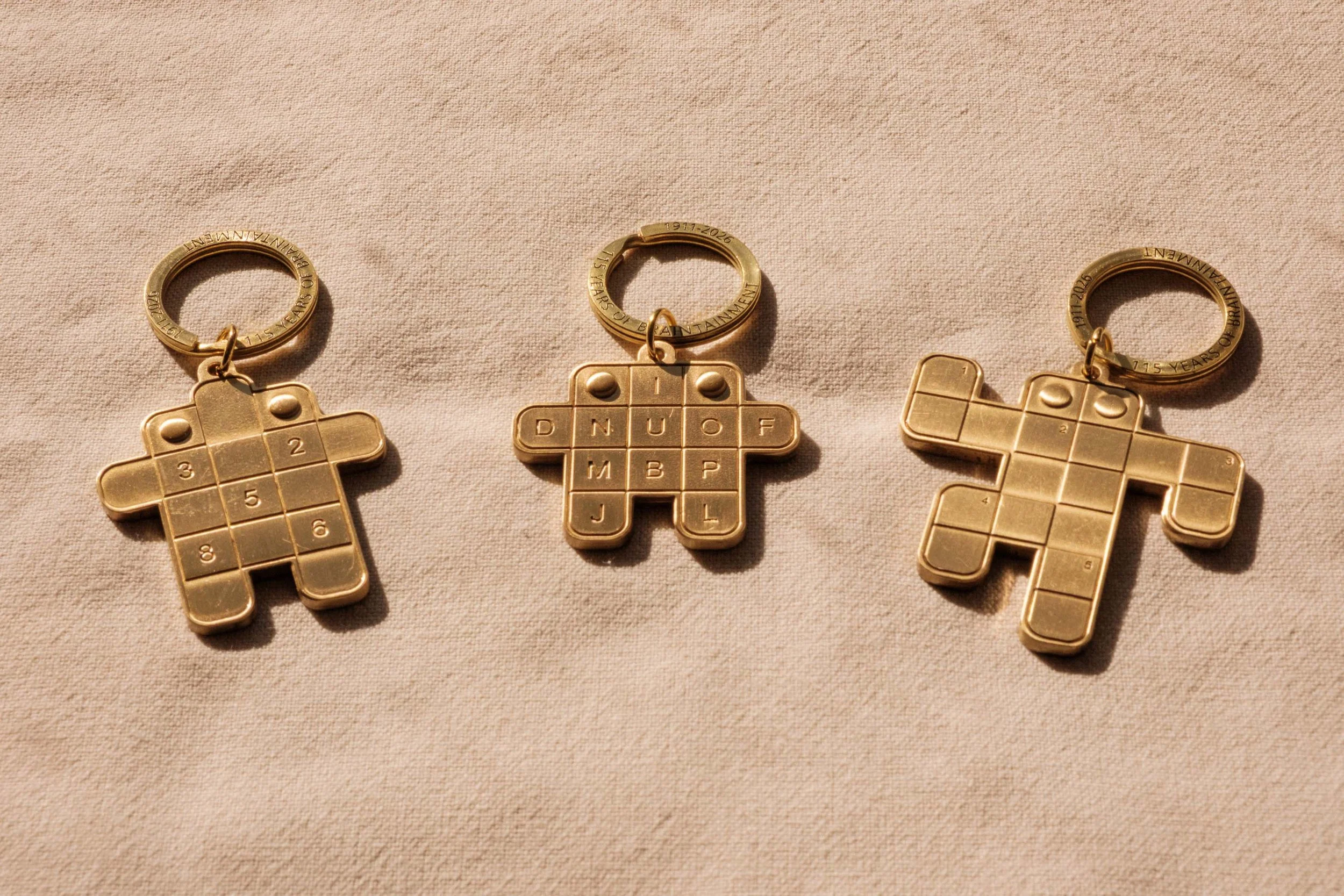 Three brass keychains shaped like tic-tac-toe boards, each with a ring at the top and engraved with years, on a beige fabric background.
