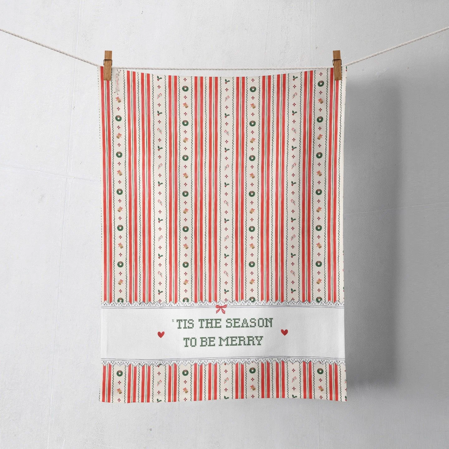 Christmas tea towel with red and green candy cane stripes wreaths bells and holly and a lace trimmed band reading Tis the Season to Be Merry in green cross stitch lettering, hanging from a clothesline