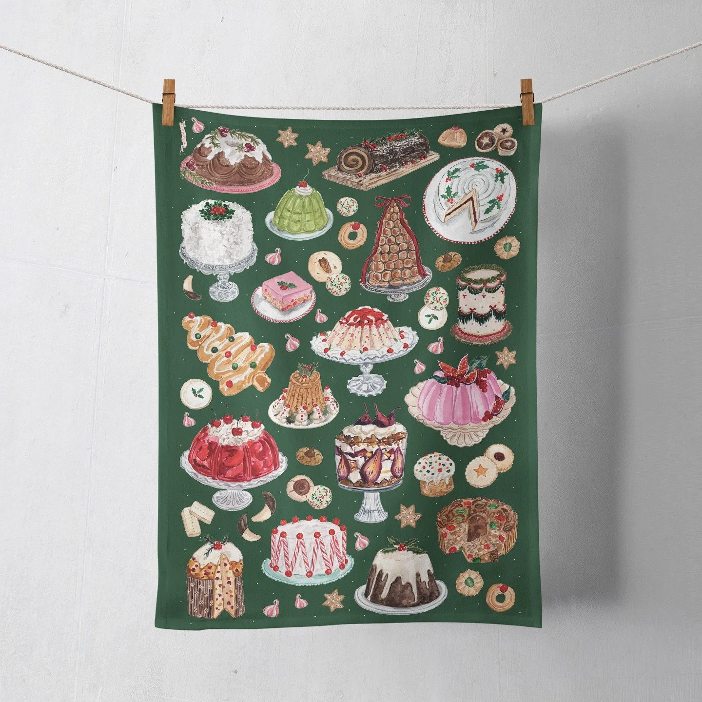 Green Christmas tea towel printed with watercolor vintage holiday desserts, including a Yule log, coconut cake, gelatin molds, croquembouche, fruitcake, trifle and Christmas cookies, hanging from a cl